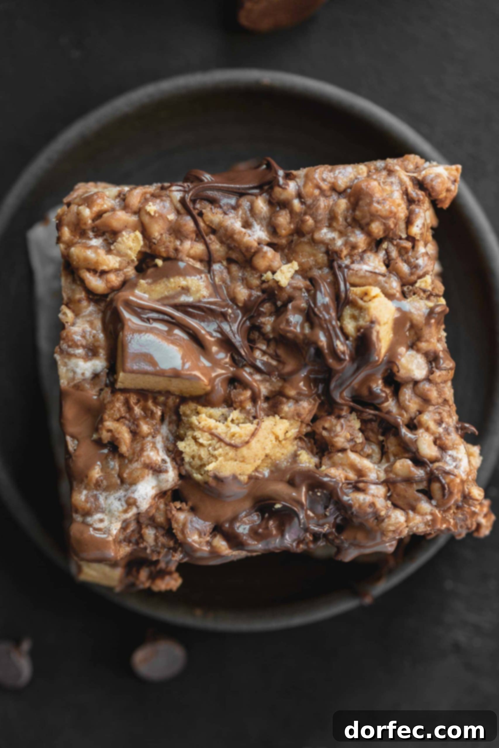 A close-up photo of a single peanut butter Rice Krispies Treat, generously topped with chopped Reese's Peanut Butter Cups and drizzled with melted chocolate chips, presented on a dark gray plate.