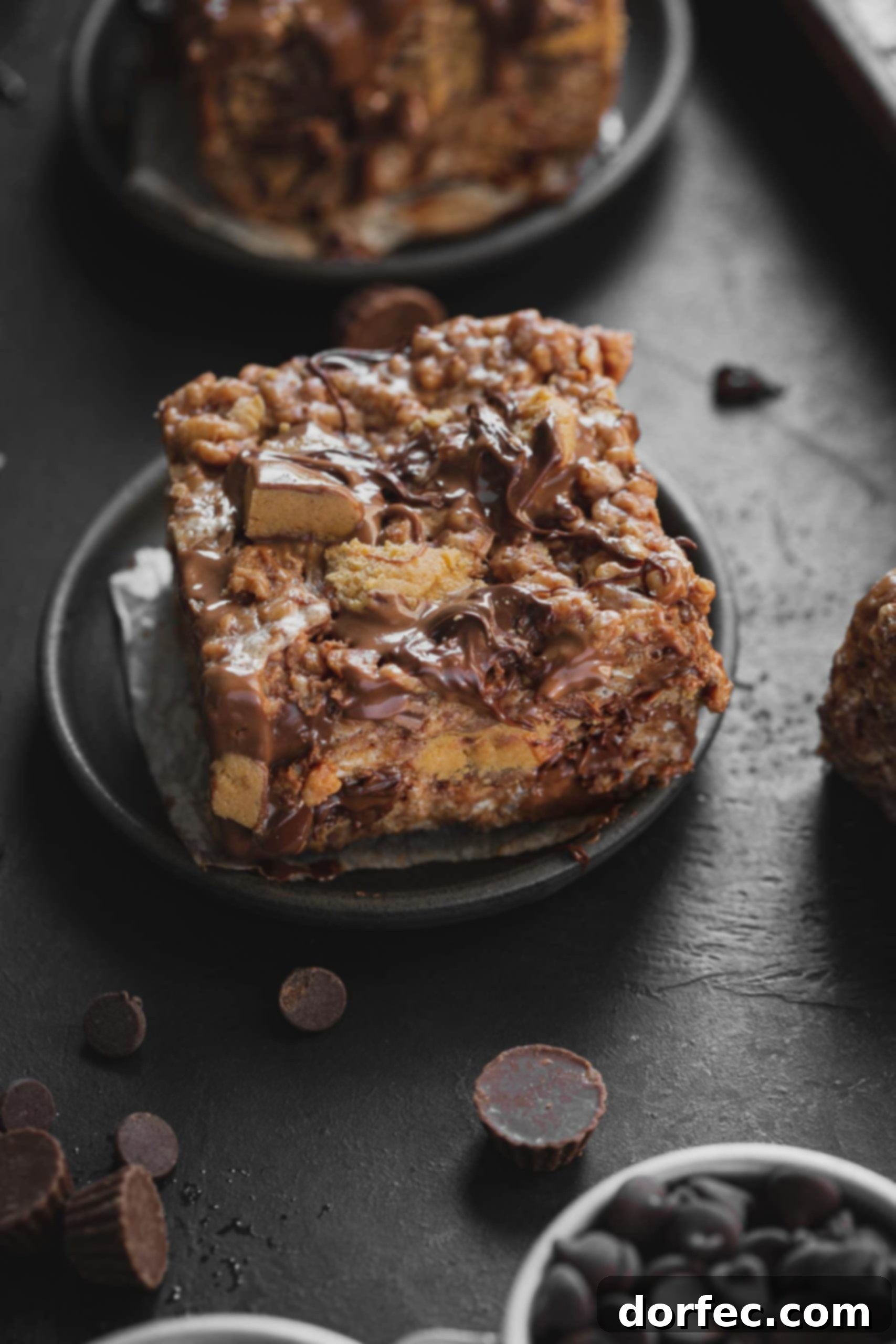 A close-up photo of a single, delectable peanut butter Rice Krispies Treat, generously loaded with chunky Reese's Peanut Butter Cups and rich, melted chocolate chips, resting on a small dark gray plate. Another similar treat is visible, slightly out of focus, in the background.