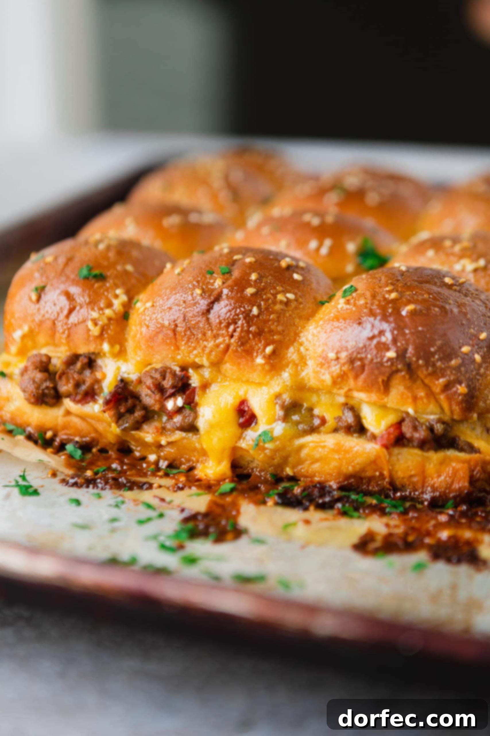A panful of cheeseburger sliders with melted cheese oozing from the rolls and visible ground beef and chunks of tomatoes.
