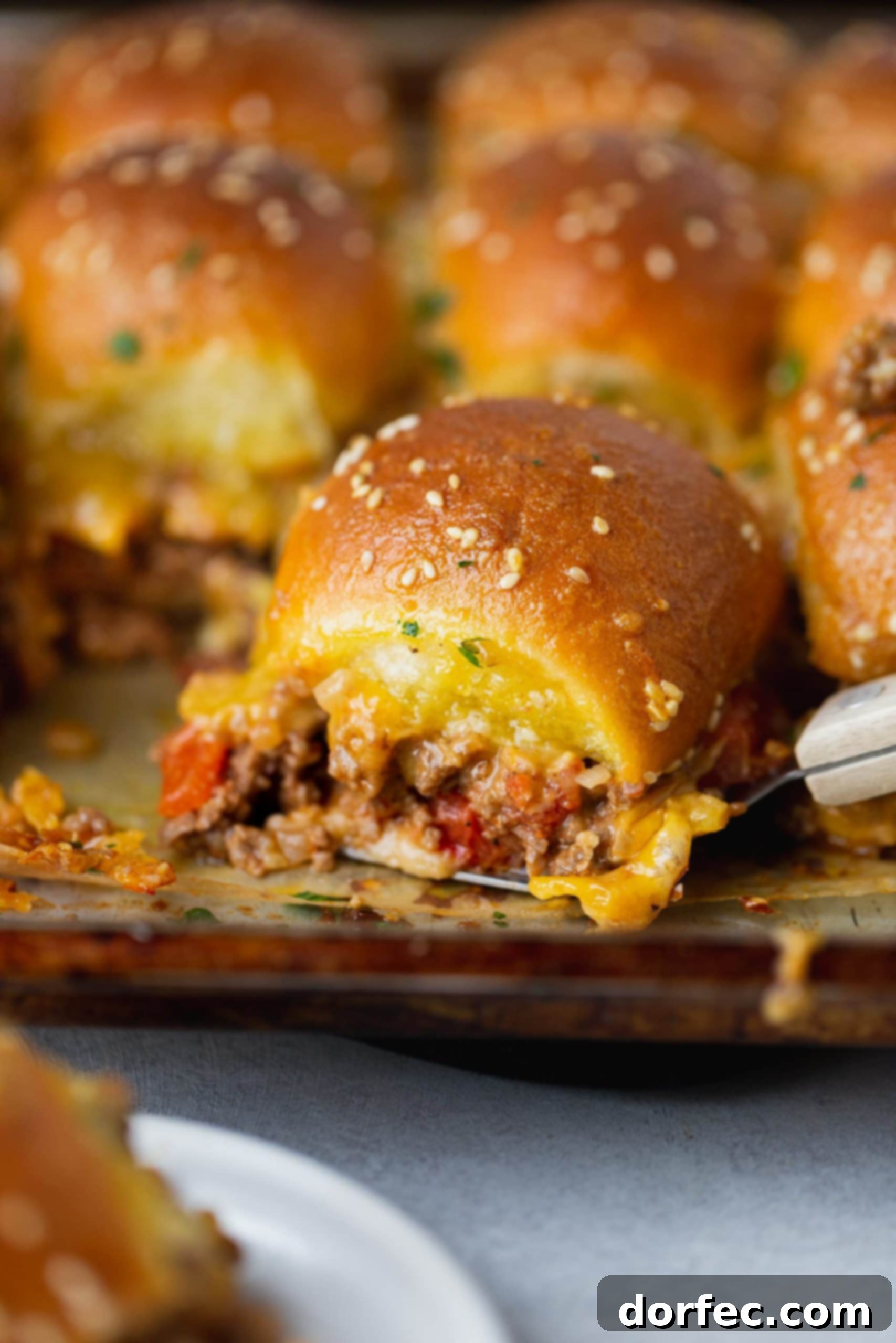 A cheeseburger slider filled with ground beef, melty cheese, and chopped tomatoes, with other sliders in the background.