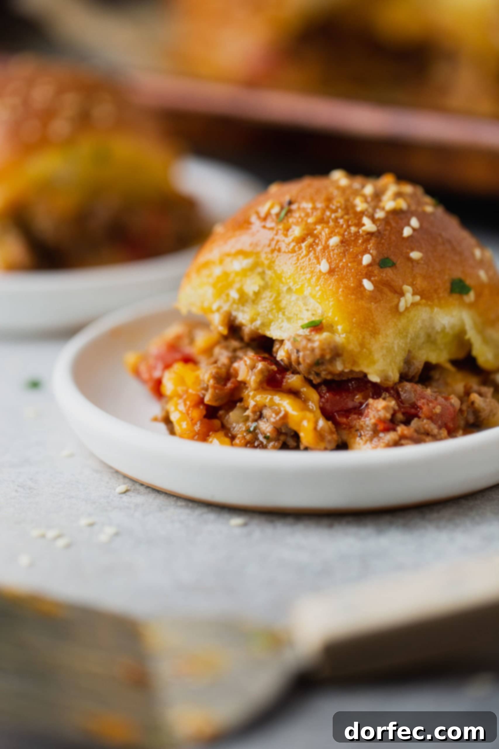 A cheeseburger slider on a plate, with golden roll packed with ground beef, melted cheese, and chopped tomatoes.