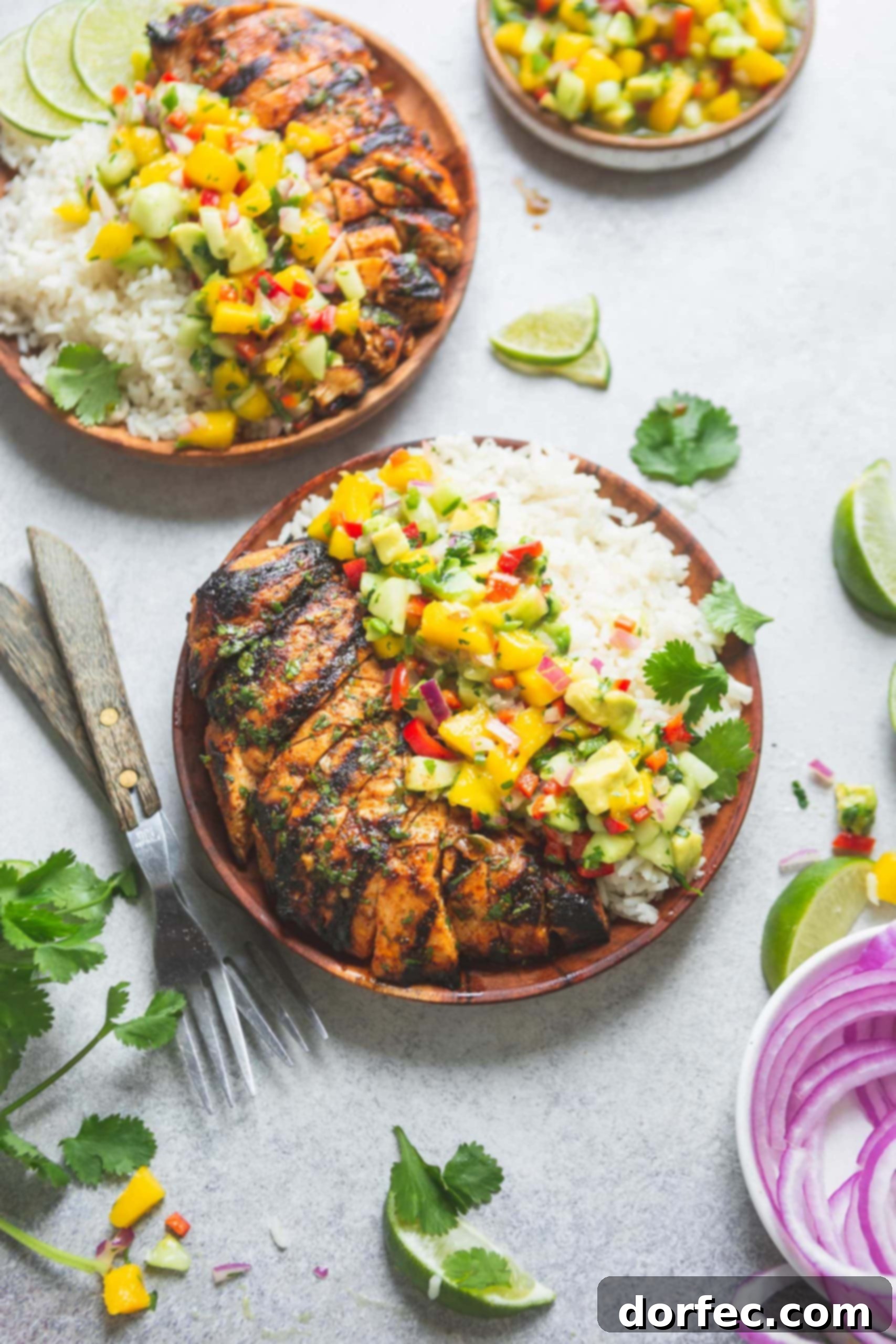 Two dinner plates filled with grilled chicken, coconut rice and mango salsa. The salsa is bright and colorful with chunks of mango and onion, peppers and cilantro. There are sliced red onions, lime wedges and forks on the table.