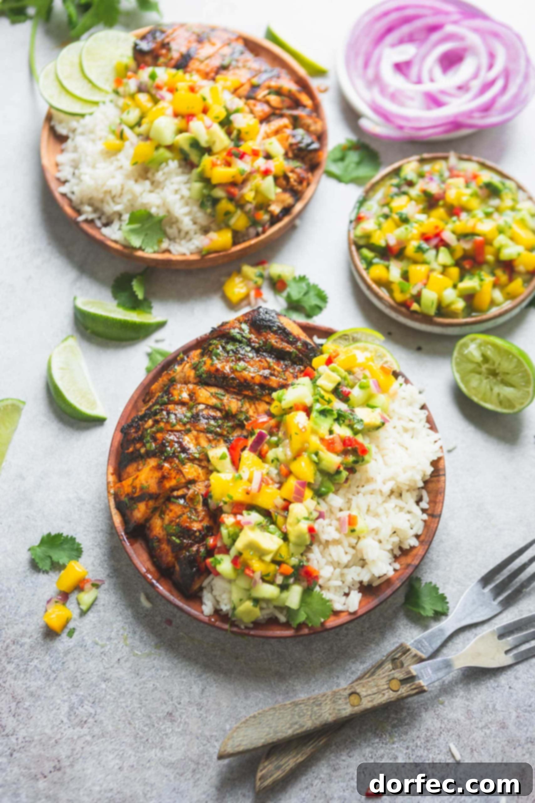 A dinner plate heaped with grilled chicken and coconut rice that is topped off with bright mango salsa. There are chunks of mango, peppers and cilantro in the salsa. A dish of salsa and another plate of chicken, rice and salsa are in the background. Wedges of lime are scattered on the table.