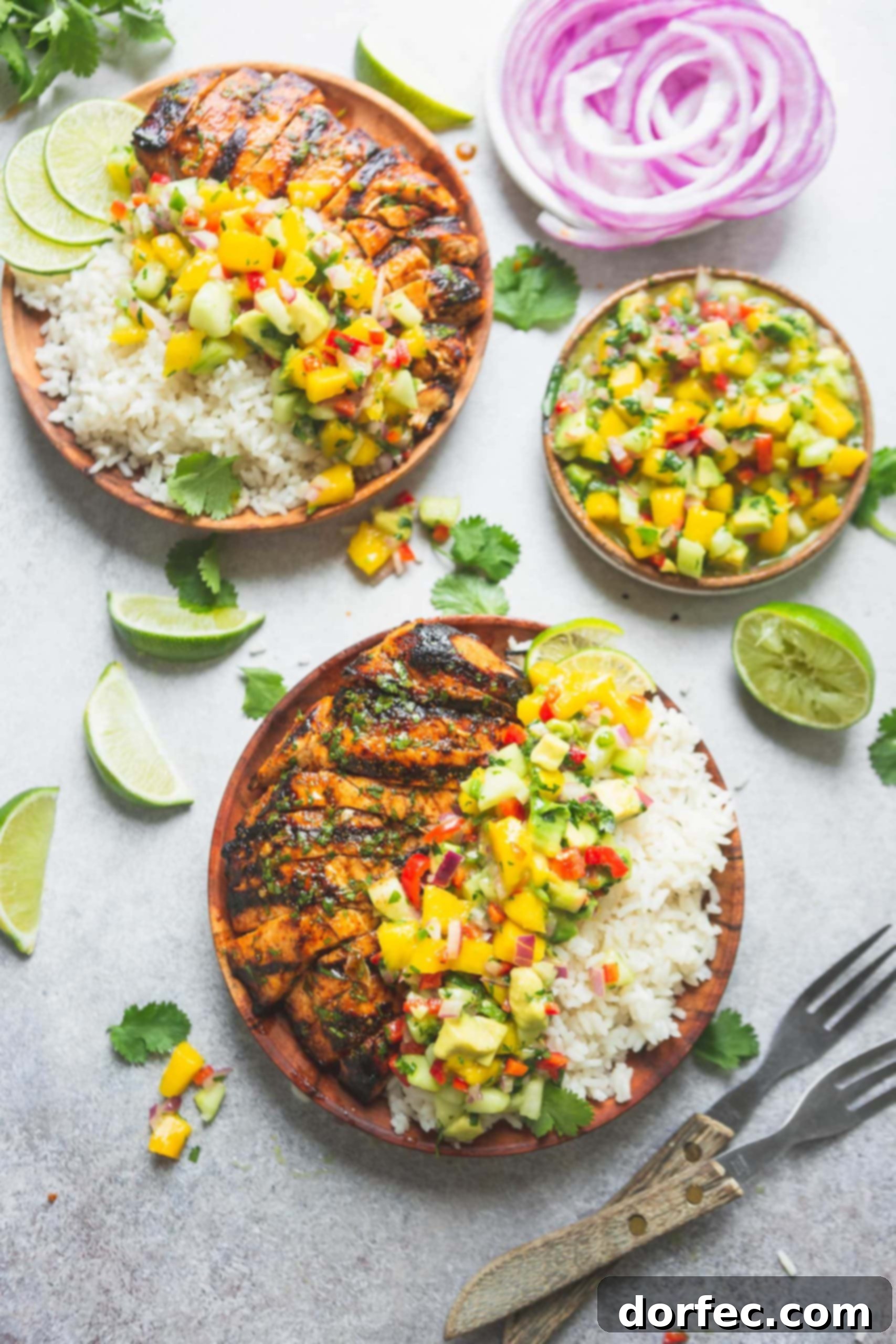 Two dinner plates piled high with grilled chicken, coconut rice and mango salsa. The salsa has chunks of mango, peppers and cilantro. A bowl of salsa in on the table and lime wedges and cilantro leaves are scattered around. There are two forks on the table too.