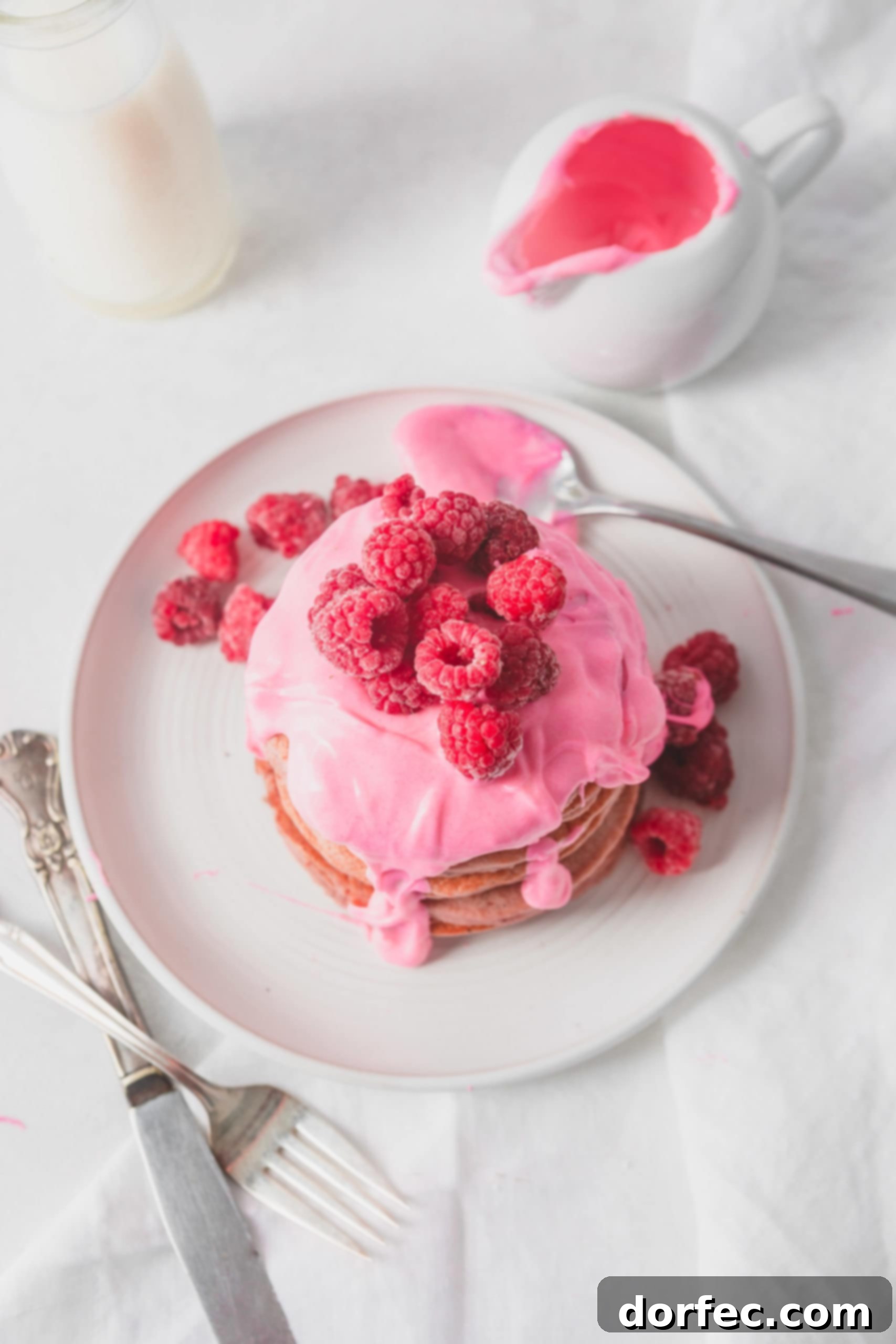 Healthy Pink Pitaya Blender Pancakes An overhead view of a stack of pink pitaya pancakes on a white plate. The pancakes are covered with a thick pink chocolate sauce topped with fresh raspberries. There is a spoon full of pink sauce on the plate and a container full of sauce in the background.