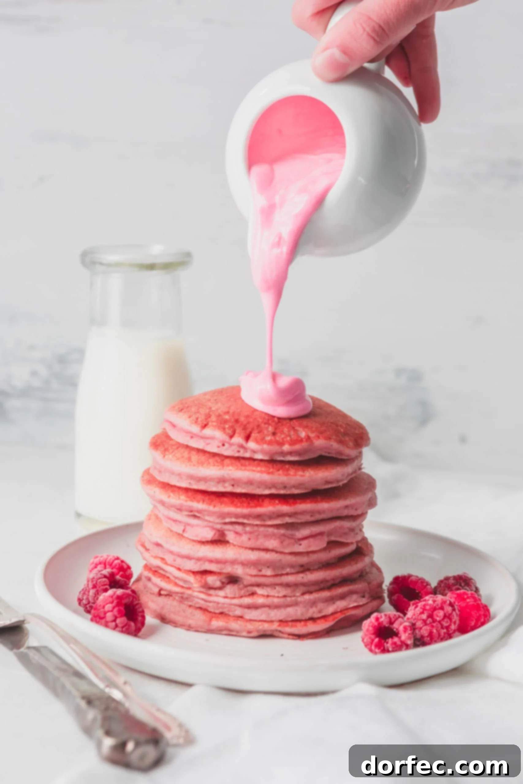 Healthy Pink Pitaya Blender Pancakes A stack of pink pancakes with a thick pink chocolate sauce being poured over the top. There are fresh raspberries on the plate with the pancakes and a container of milk in the background. with