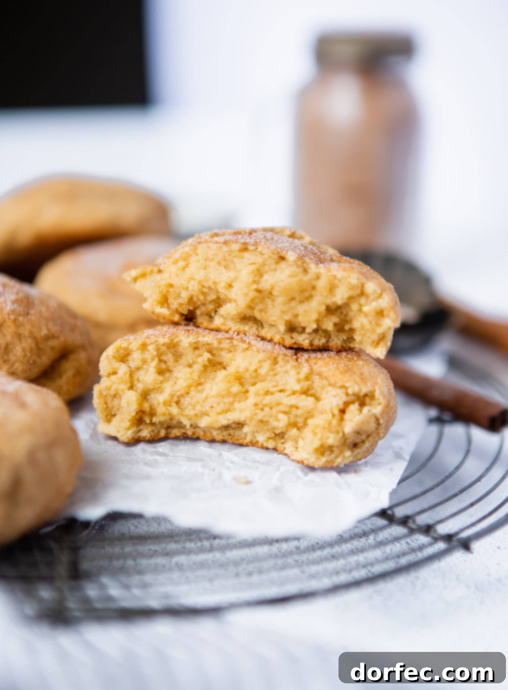 Thick Snickerdoodle Cookies Snickerdoodles on a cooling rack. One cookies is broken in half exposing the soft crumbly inside.