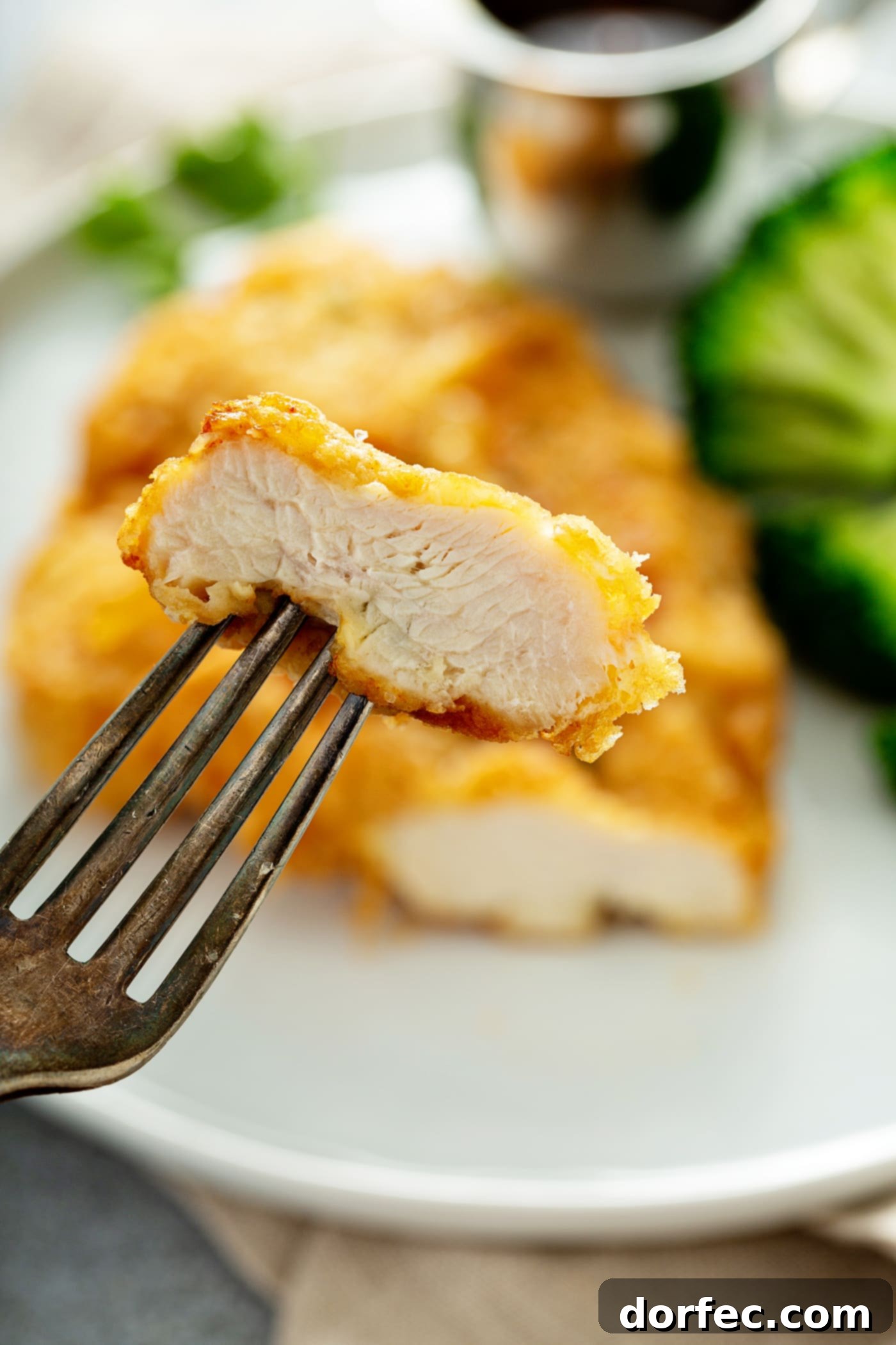 Authentic Southern Crispy Fried Chicken 3 A golden brown fried chicken breast with a slice cut off and held up in the tines of a fork. There is broccoli beside the chicken on the plate.