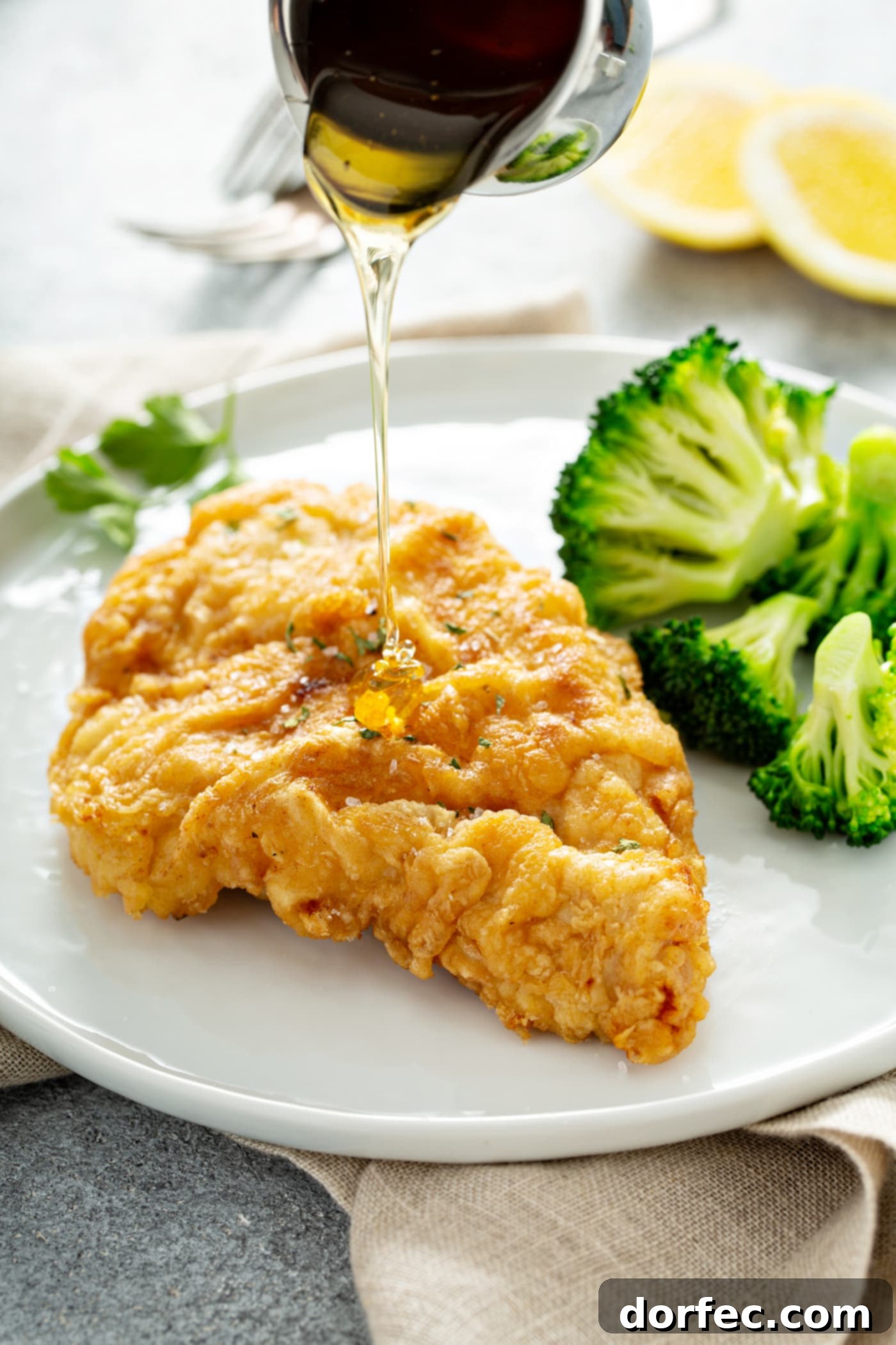Authentic Southern Crispy Fried Chicken 4 A chicken breast that has been fried to a golden brown, resting on a white dinner plate. There is steamed broccoli next to the chicken, and a sauce is being drizzled over the chicken.