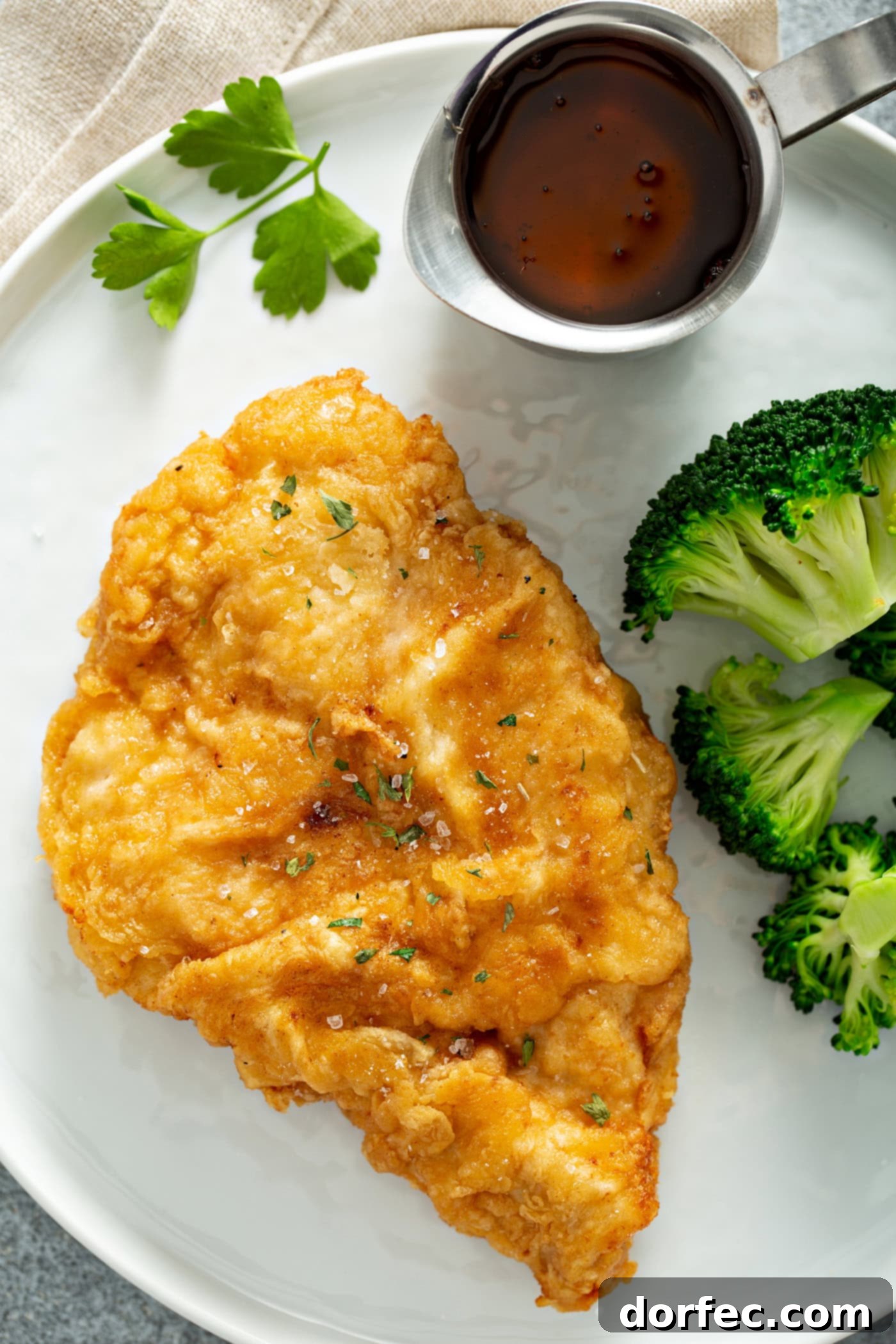 Authentic Southern Crispy Fried Chicken 5 A chicken breast that has been fried to a golden brown, resting on a white dinner plate. There is also steamed broccoli next to the chicken and a small pitcher of sauce.