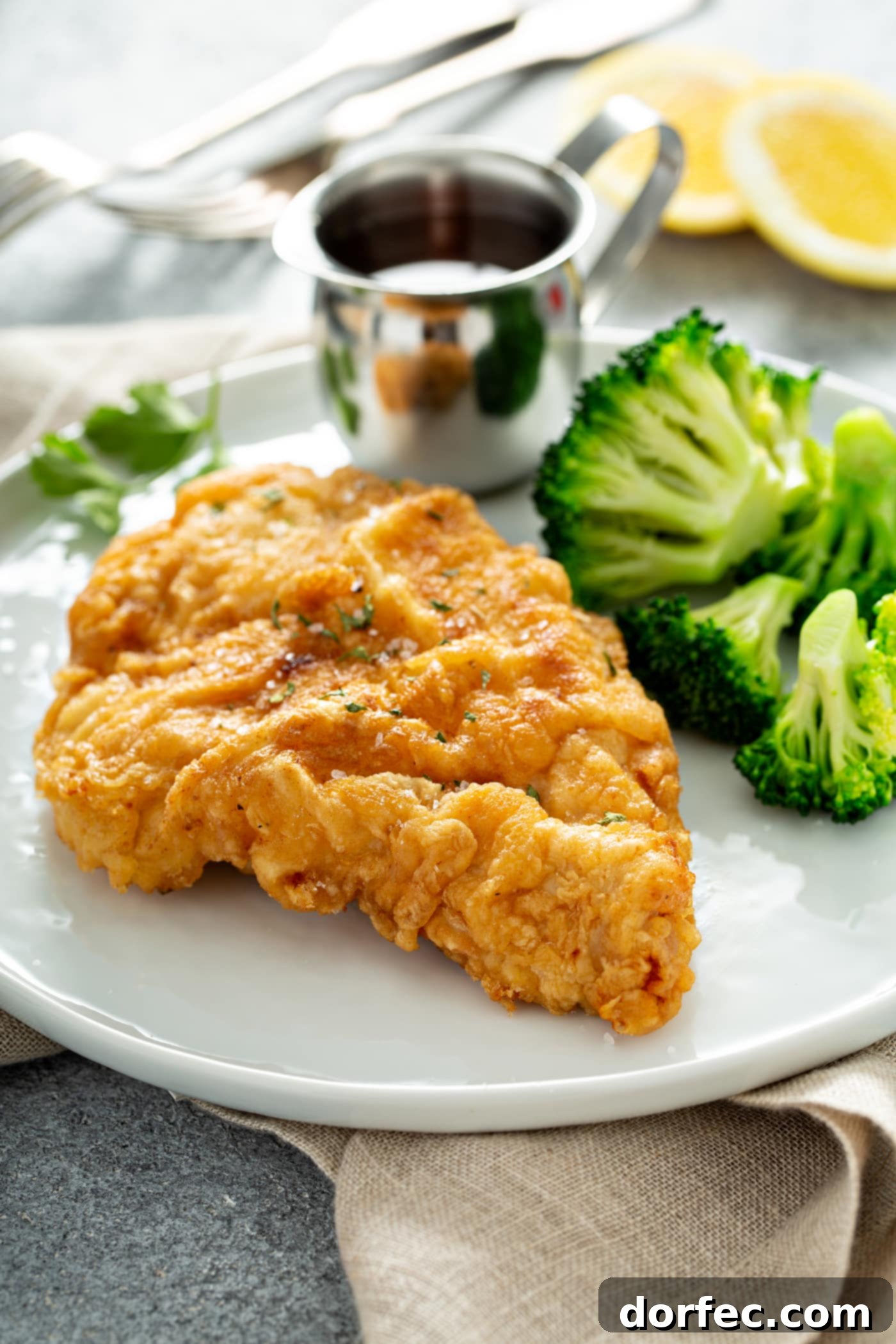 Authentic Southern Crispy Fried Chicken 6 A piece of chicken fried to a perfect golden brown. There is a silver pitcher of sauce and steamed broccoli next to the chicken on a white dinner plate.