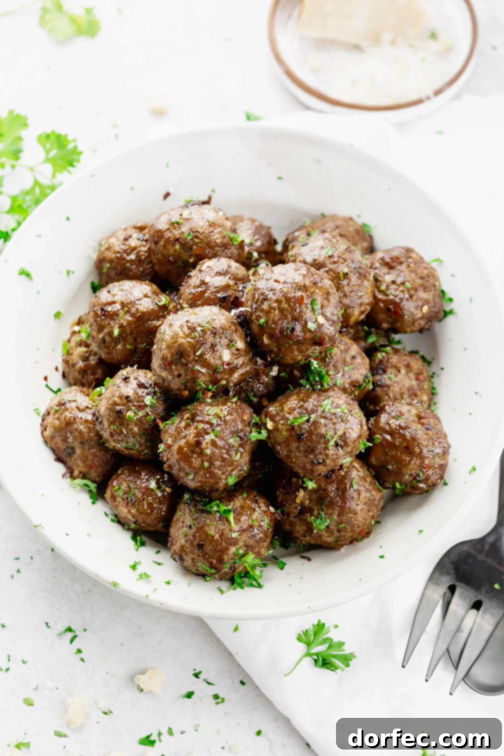 A large, rustic bowl brimming with freshly cooked meatballs, generously garnished with chopped parsley. A fork and spoon are placed elegantly next to the bowl.