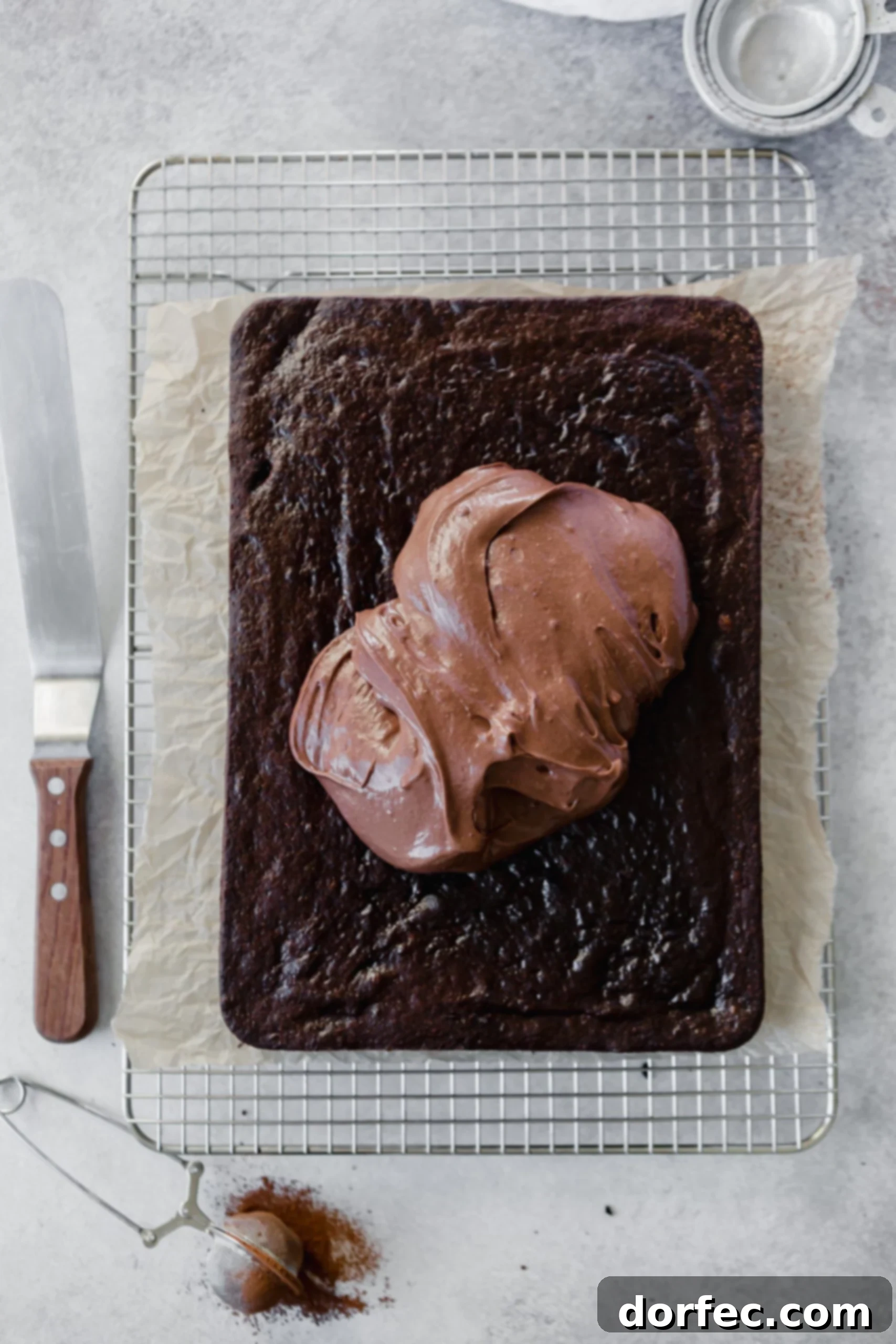 Chocolate Mayonnaise Cake A 9x13 inch chocolate mayonnaise cake on a cooling rack, with a mound of rich chocolate cream cheese frosting waiting to be spread. A frosting spatula rests beside the cake, ready for action.