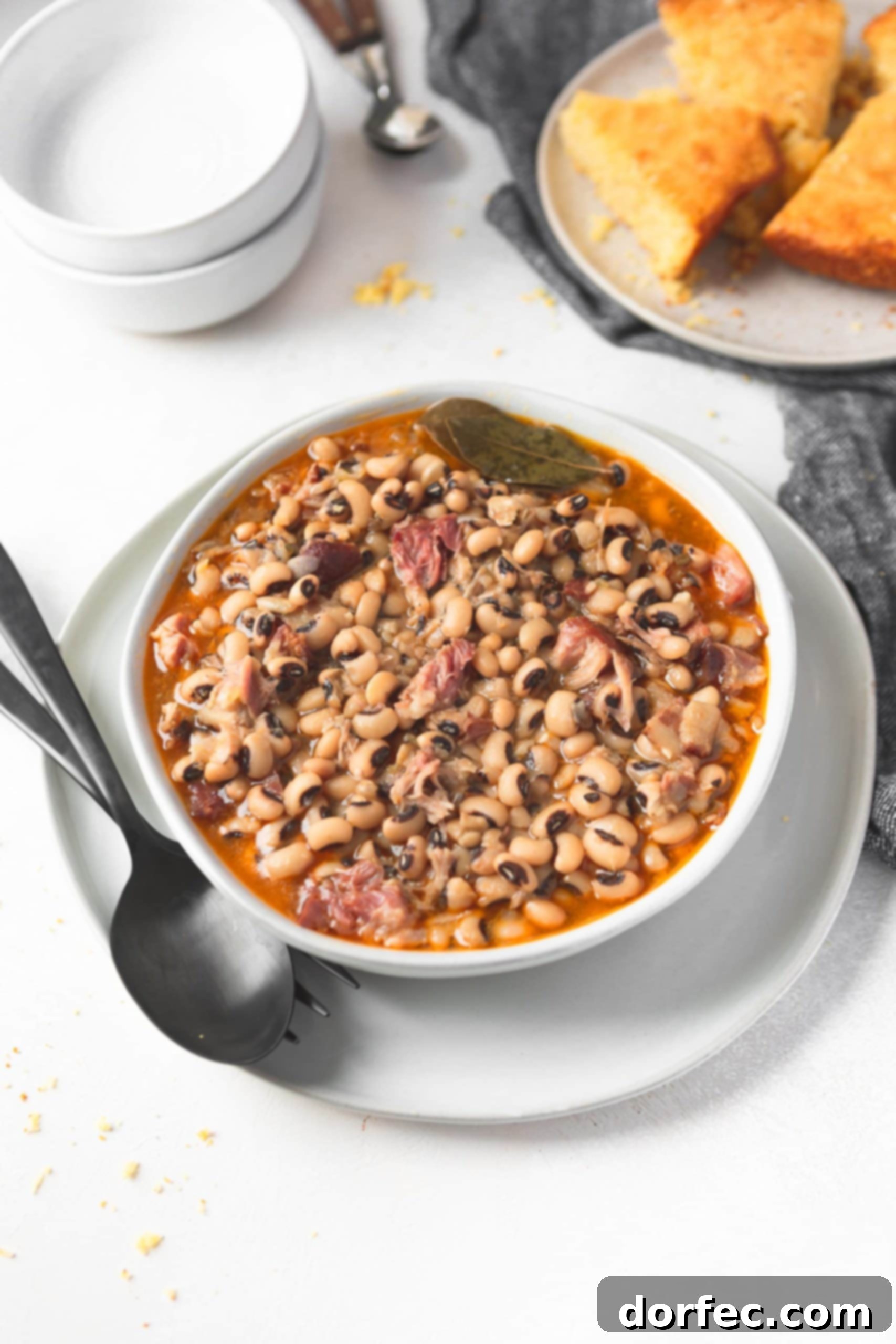 A picture of a bowl of southern black eyed peas, garnished and ready to be served. The bowl is sitting on a saucer, accompanied by a fork and spoon. A plate of golden cornbread is blurred in the background, suggesting a complete Southern meal. The overall scene evokes warmth and comfort.