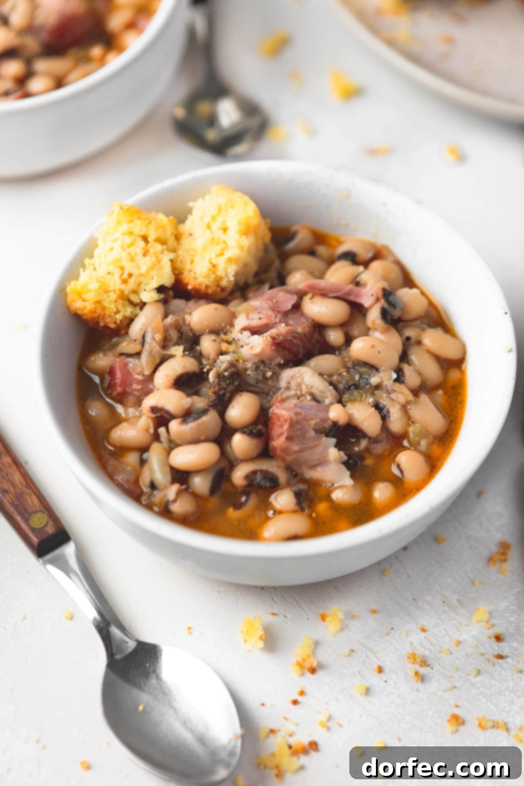 A hearty bowl of Southern black eyed peas, generously studded with tender chunks of ham. A broken piece of golden cornbread rests invitingly on the side, ready for dipping. The focus is on the rich texture and appealing presentation of the meal.