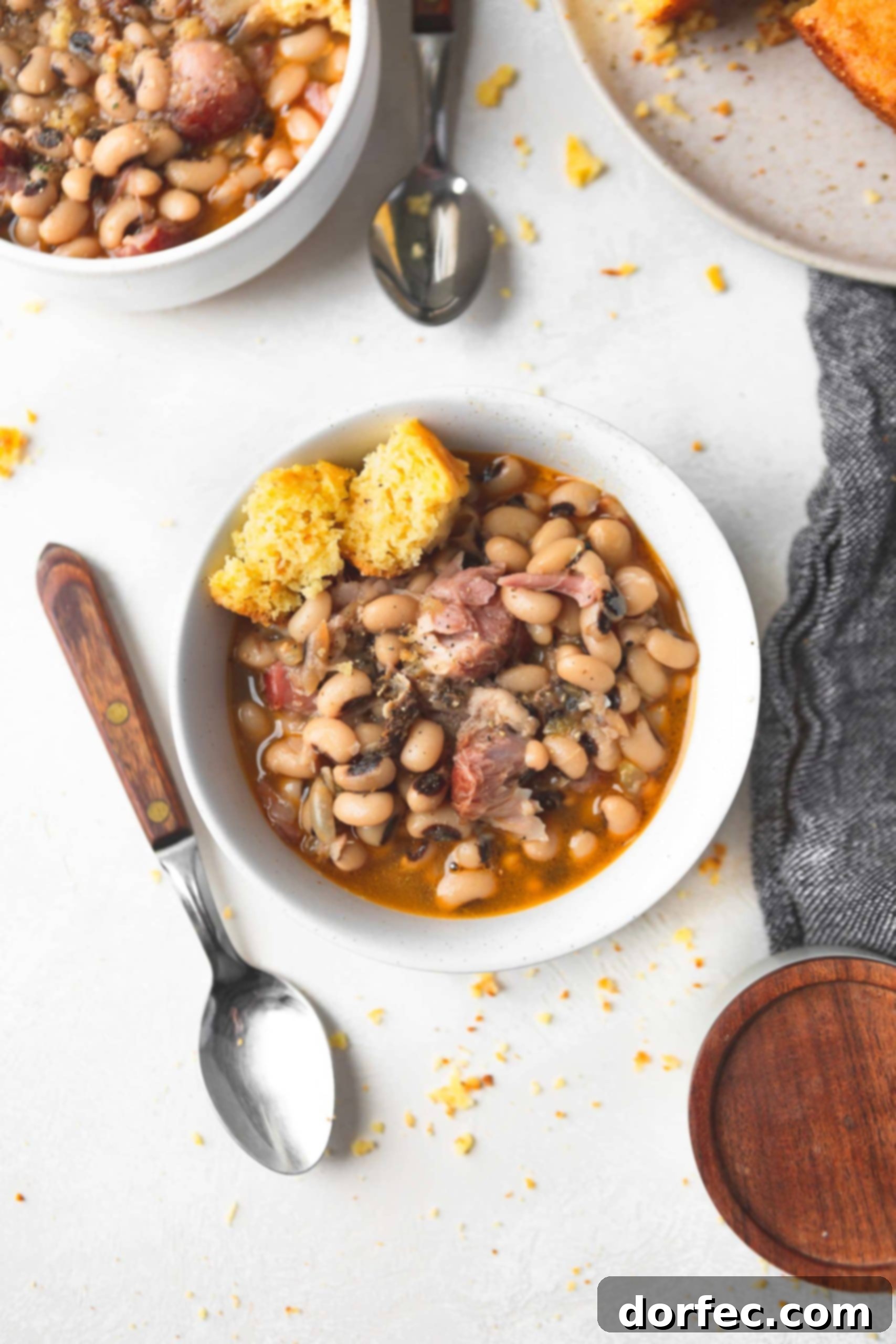 A inviting bowl of southern black eyed peas, brimming with savory chunks of ham and a rustic piece of cornbread for dipping. A spoon rests beside the bowl, emphasizing its readiness to be enjoyed. The image captures the essence of a comforting and traditional meal.