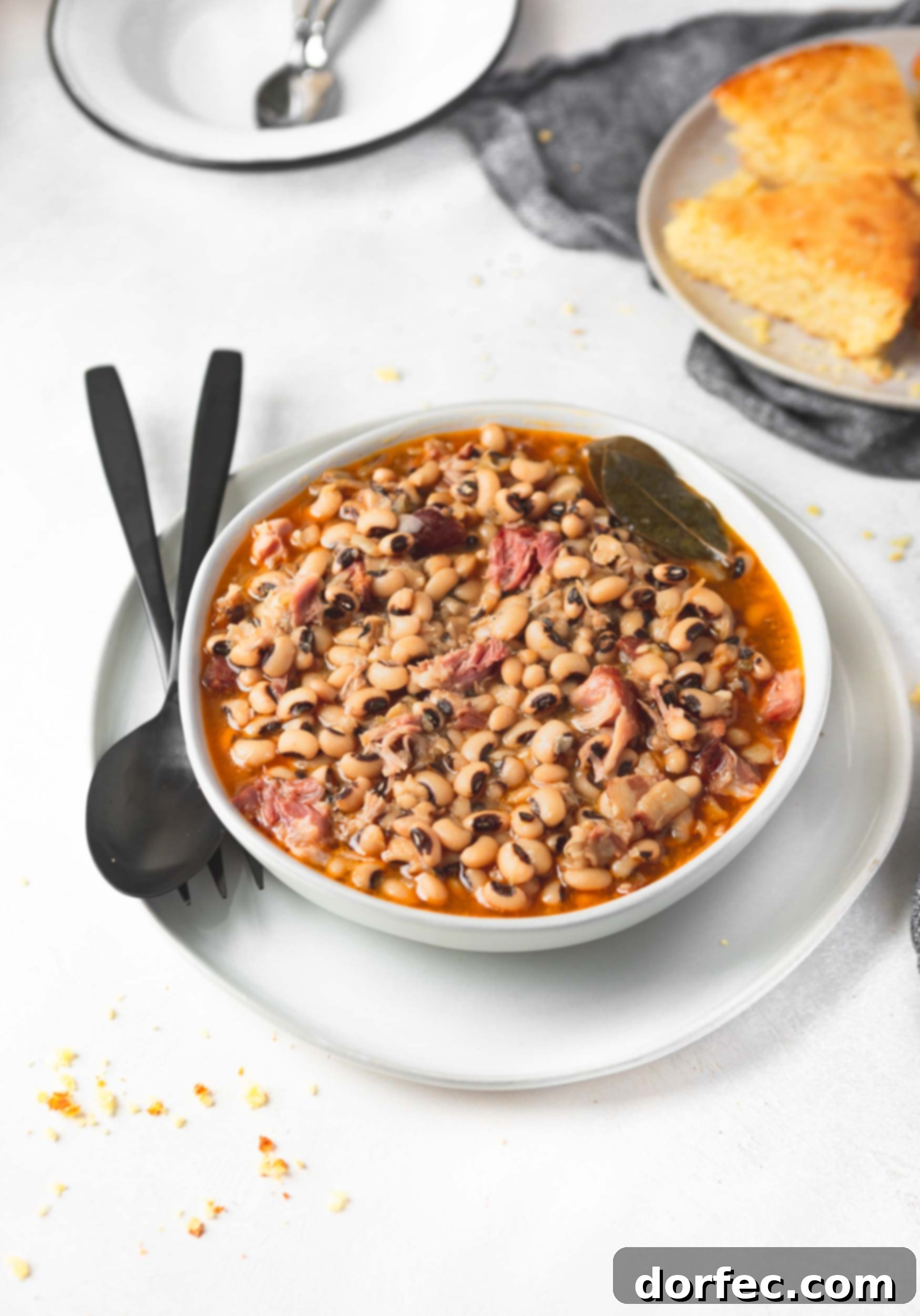 A beautifully composed shot of a bowl of black eyed peas, resting on a saucer, with a fork and spoon ready. In the soft-focus background, a plate of cornbread hints at the complete, comforting meal. The image emphasizes the inviting nature and rich texture of the dish.