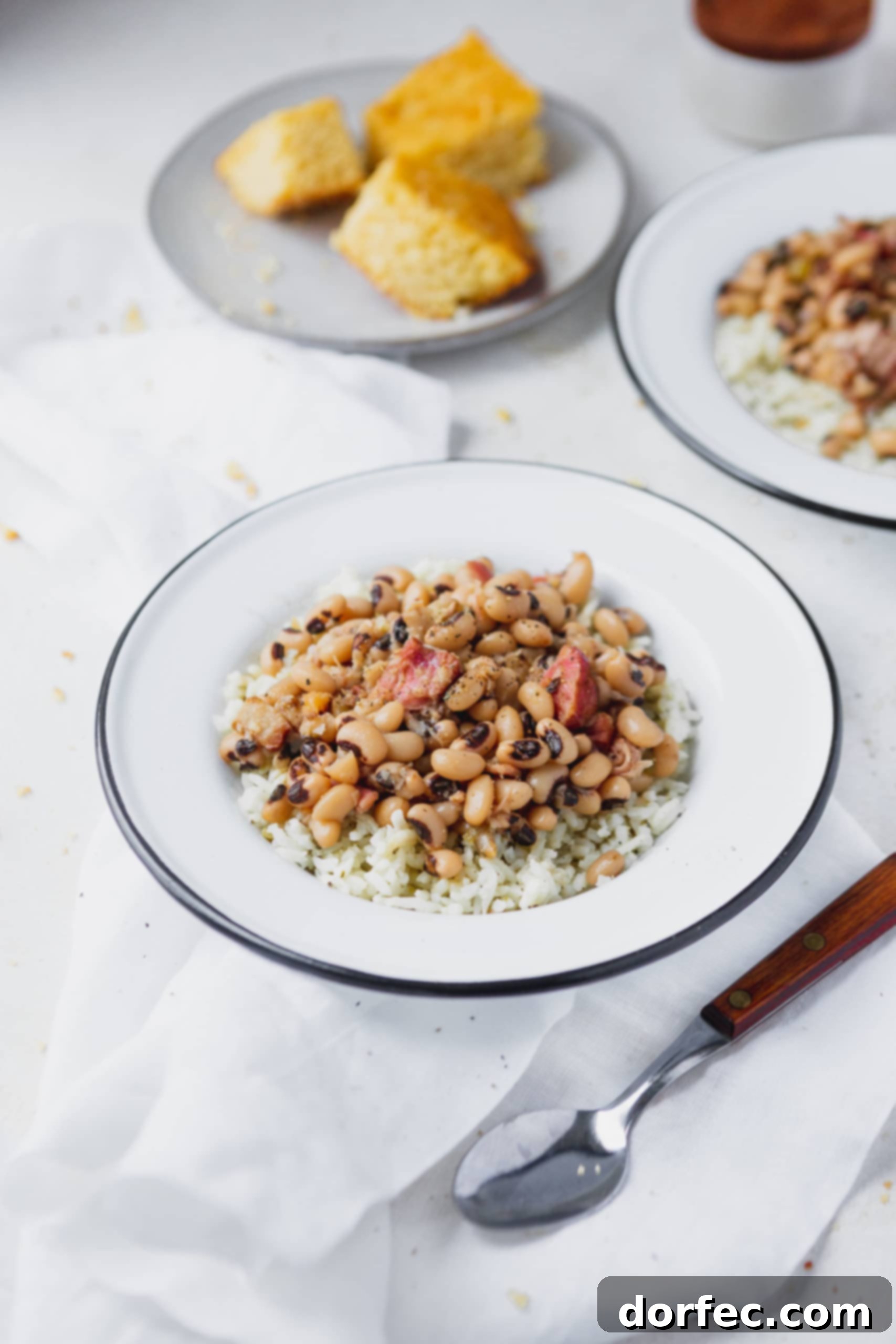 Instant Pot Black Eyed Peas A plate of rice with southern Instant Pot black eyed peas on top. There is a spoon beside the plate and another plate of peas and a plate of corn bread in the background.