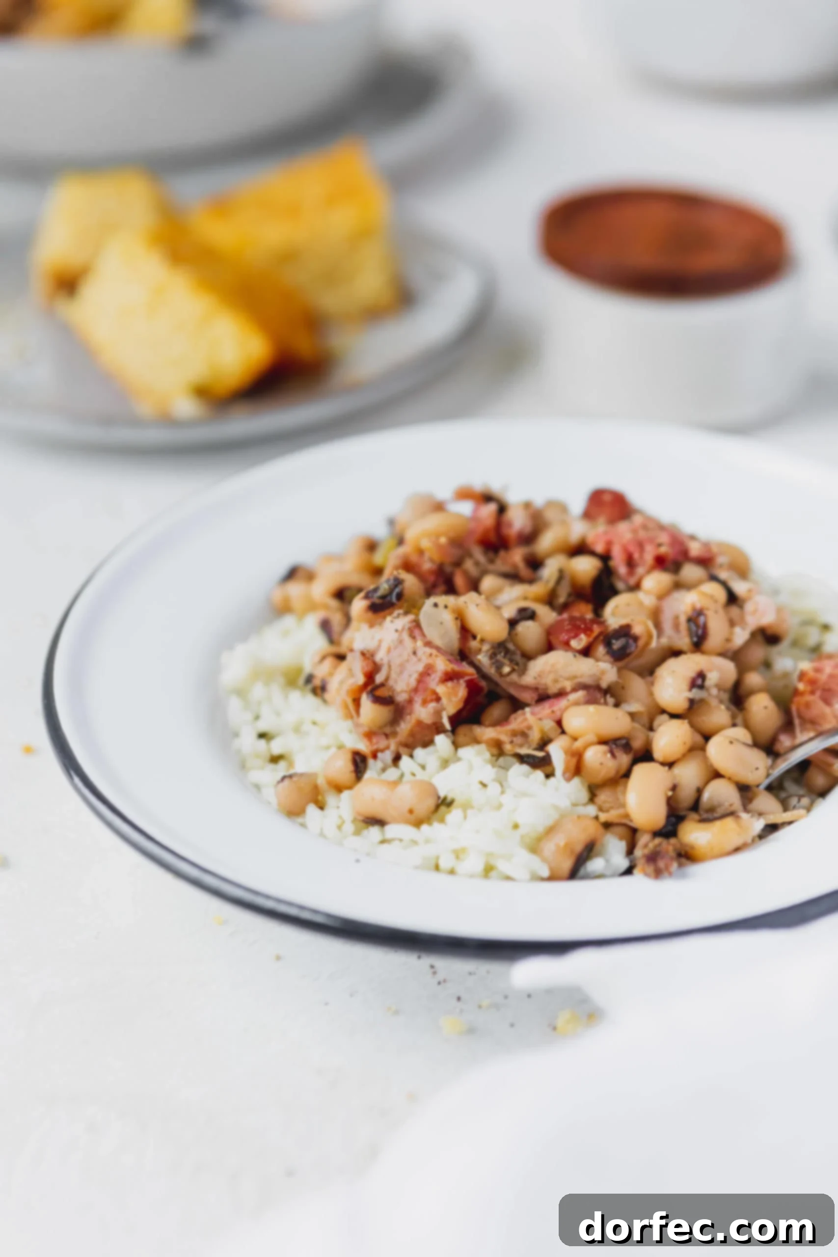 Instant Pot Black Eyed Peas A plate of southern Instant Pot black eyed peas over rice. There are chunks of ham in the peas and a plate of cornbread in the background.