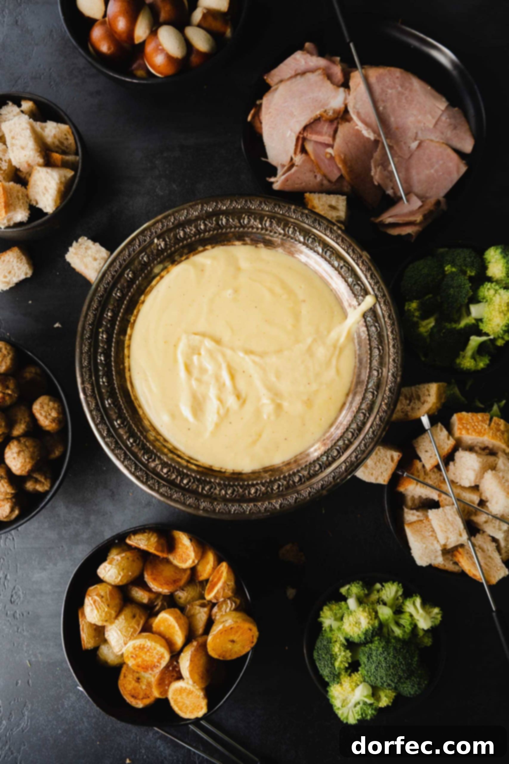 Perfect Cheese Fondue and What to Dip in Fondue A beautiful bowl of creamy cheese fondue, garnished with a sprig of rosemary. It is surrounded by an inviting spread of bread cubes, crisp apple slices, vibrant vegetables, and cured meats, all perfect for dipping.