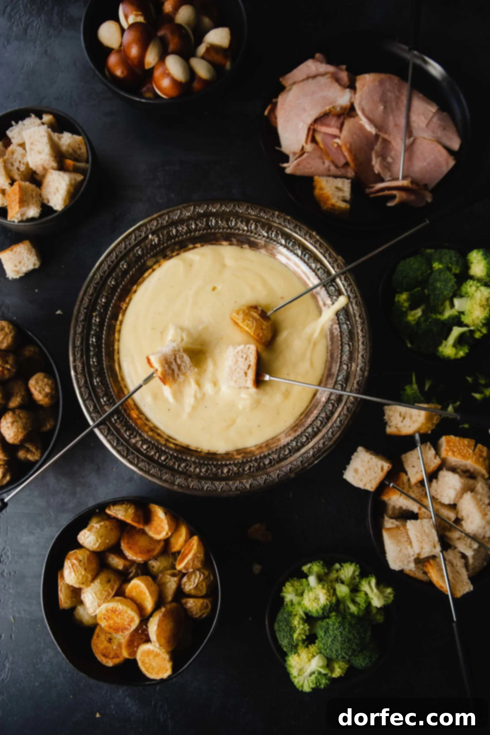 Perfect Cheese Fondue and What to Dip in Fondue A tempting bowl of cheese fondue sits as the centerpiece of a festive spread. Skewers laden with bread are being dipped, creating delightful swirls in the melted cheese. Surrounding the pot is an abundant selection of dippers: savory meats, fresh fruits, crunchy vegetables, and more bread, promising a rich and varied culinary experience.