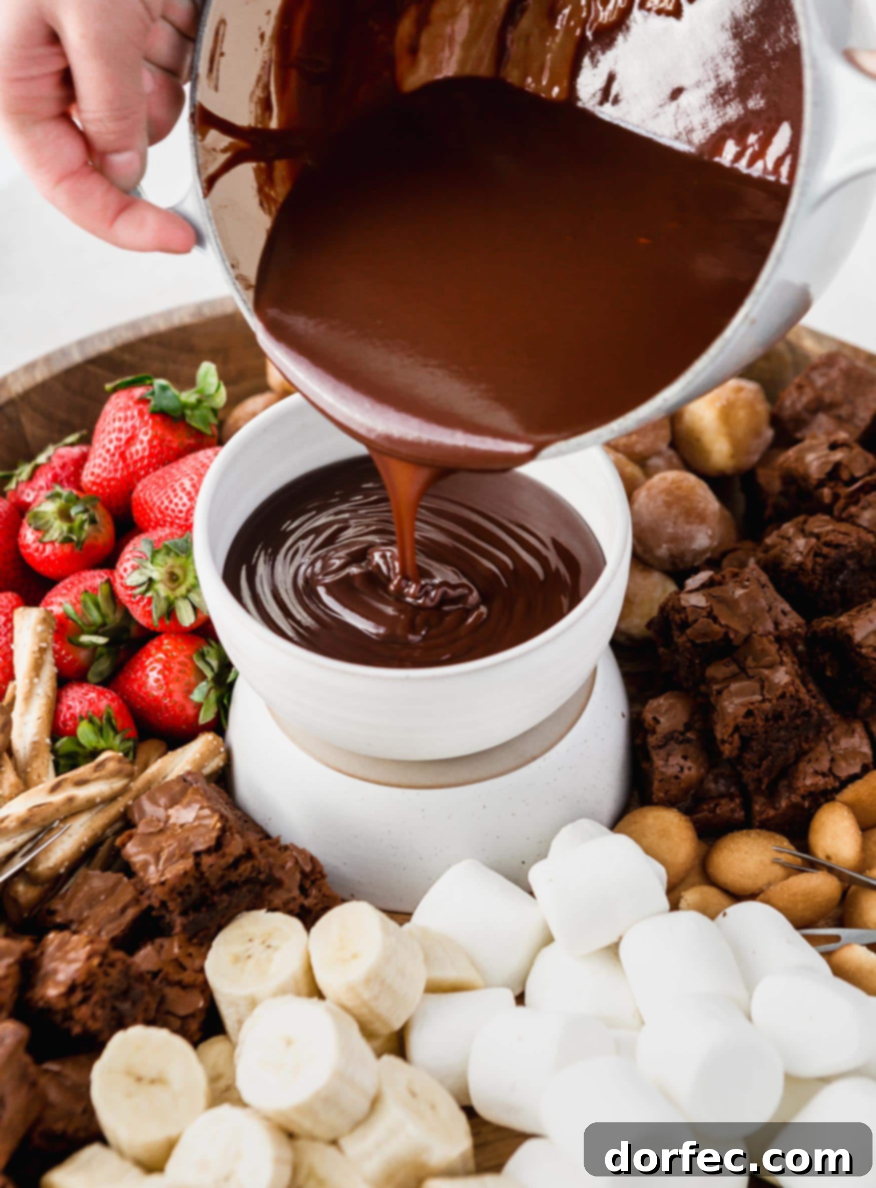 Pouring Chocolate Fondue Chocolate fondue being poured into a serving bowl. The bowl is on a tray surrounded by strawberries, marshmallows, cookies and nuts to be dipped in the fondue.