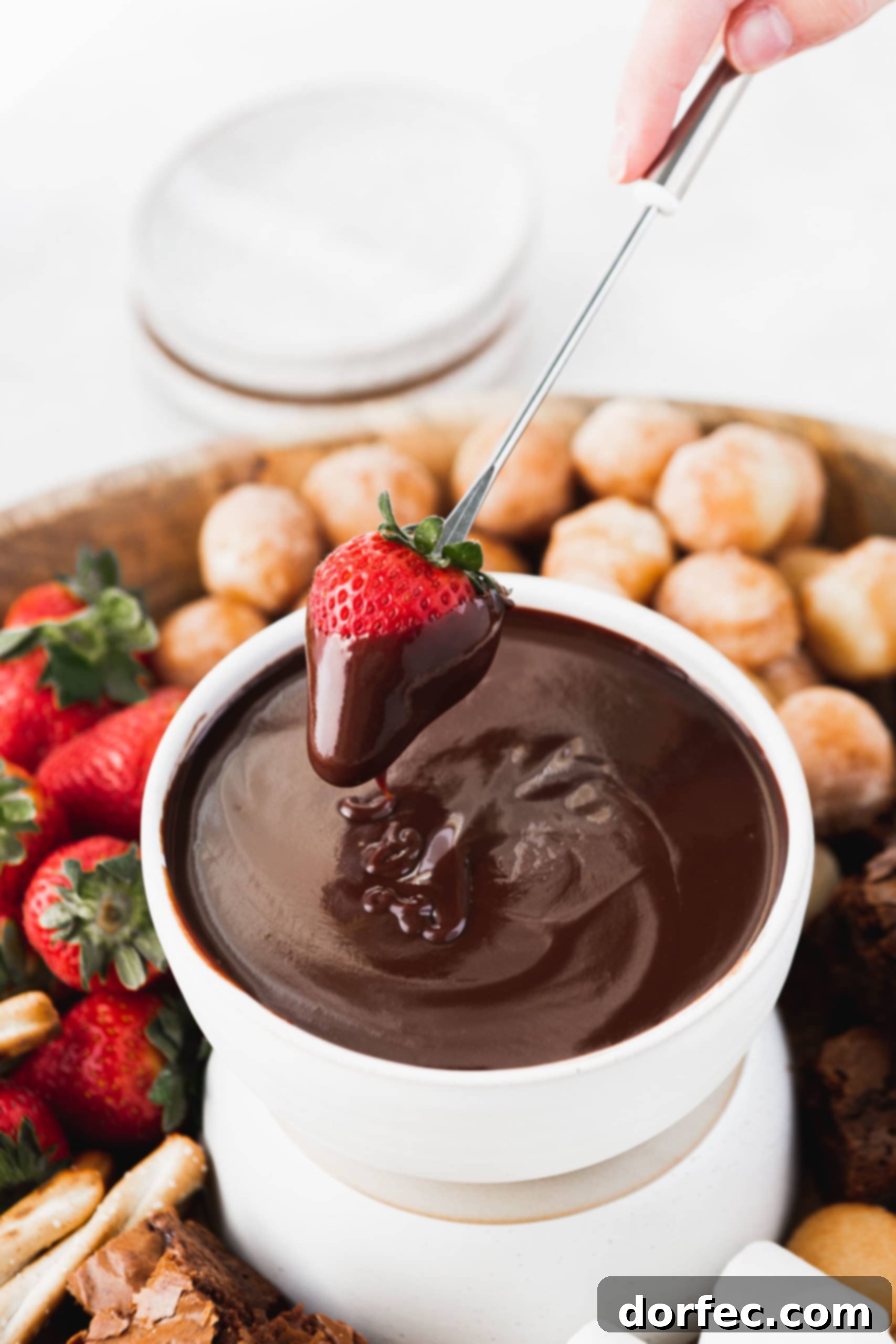Rich Chocolate Fondue A bowl of chocolate fondue into which a strawberry has been dipped. There are more strawberries and brownies and donut holes in the background.