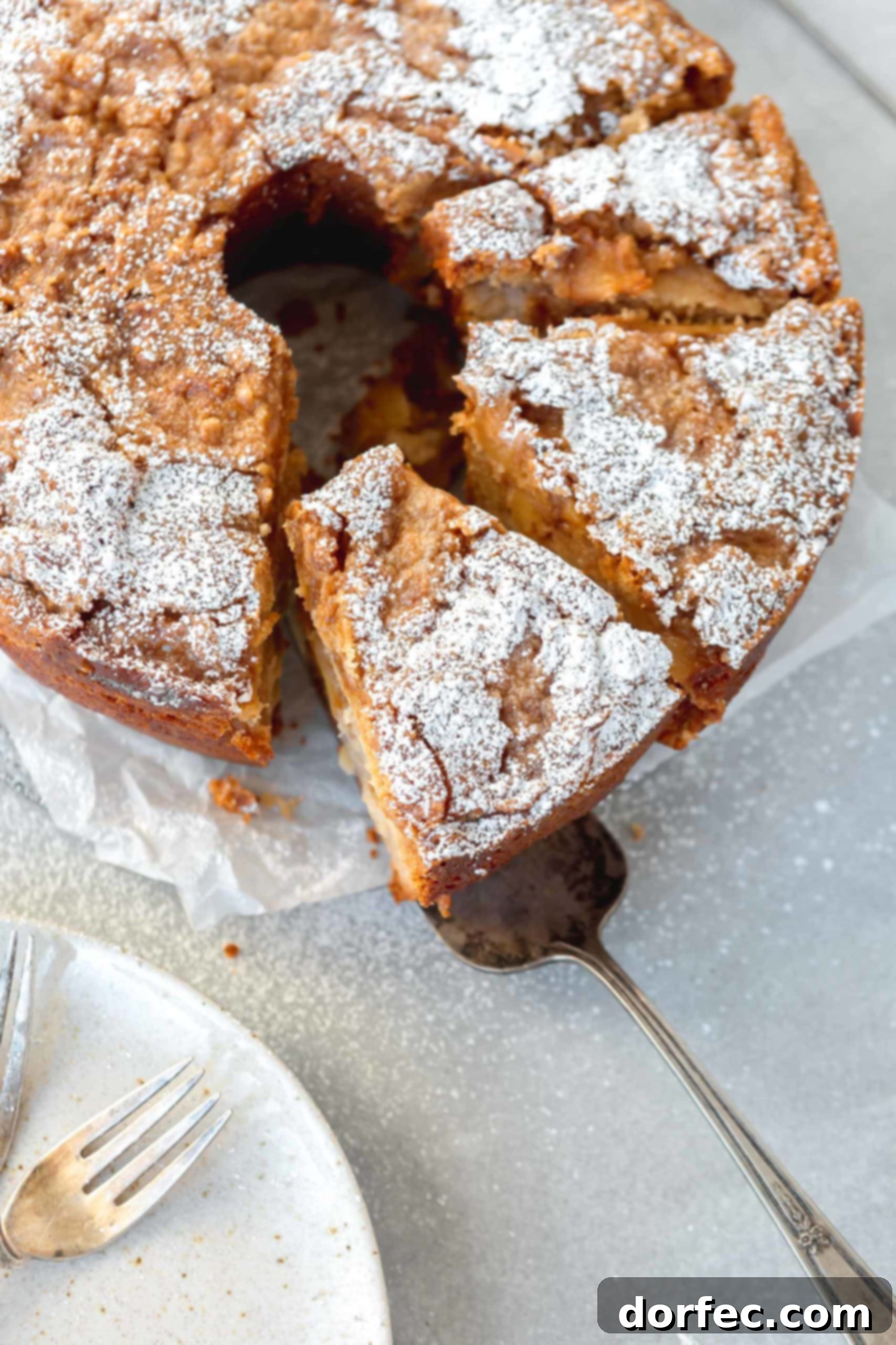 A picture of a Jewish apple cake. It is shaped like an angel food cake and has one slice cut and partially removed. The cake has powdered sugar on top.