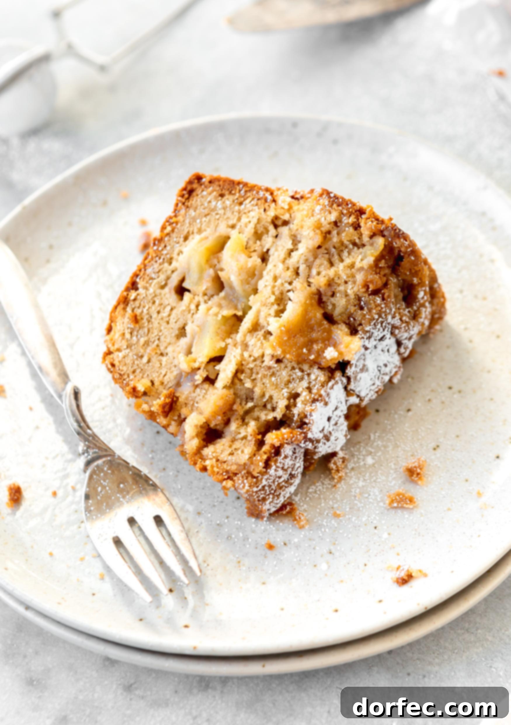 A slice of Jewish apple cake on a white dessert plate with a fork next to it. The cake is dense, and filled with apples. There is powdered sugar on the cake top.