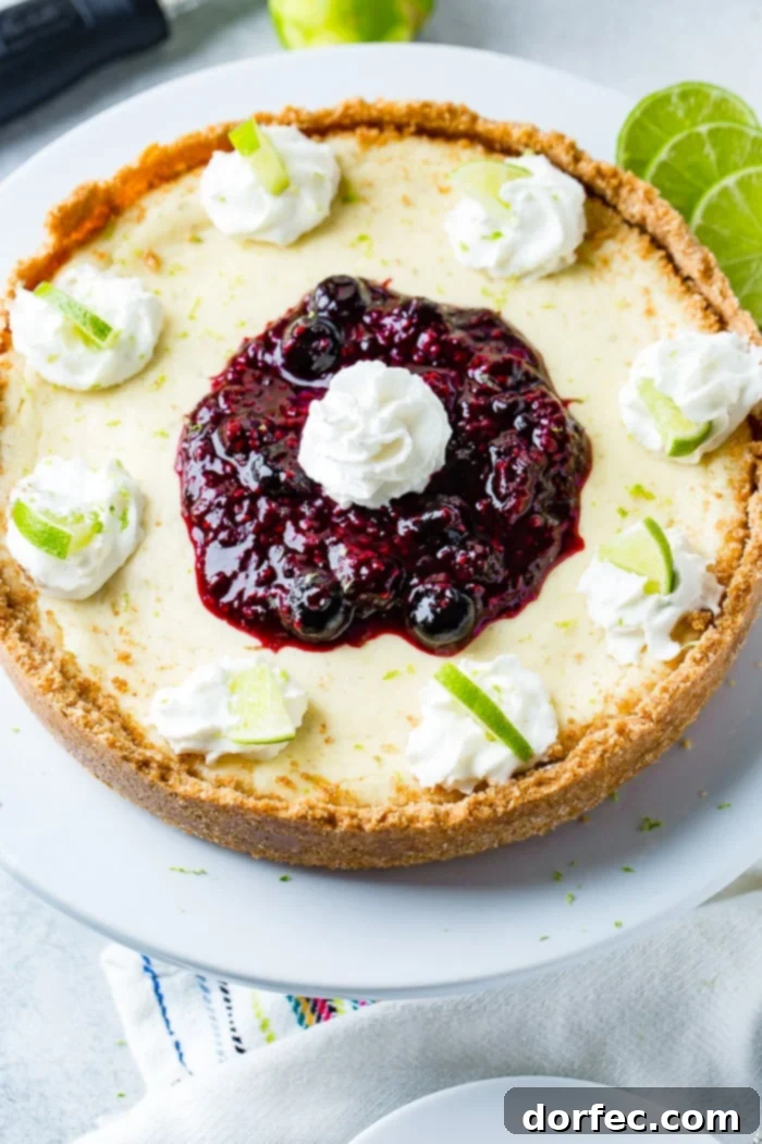 An aerial photo of a whole cheesecake on a white serving platter with dollops of whipped cream around the edges with a slice of lime in each dollop and a pile of triple berry sauce in the middle with a dollop of whipped cream on top