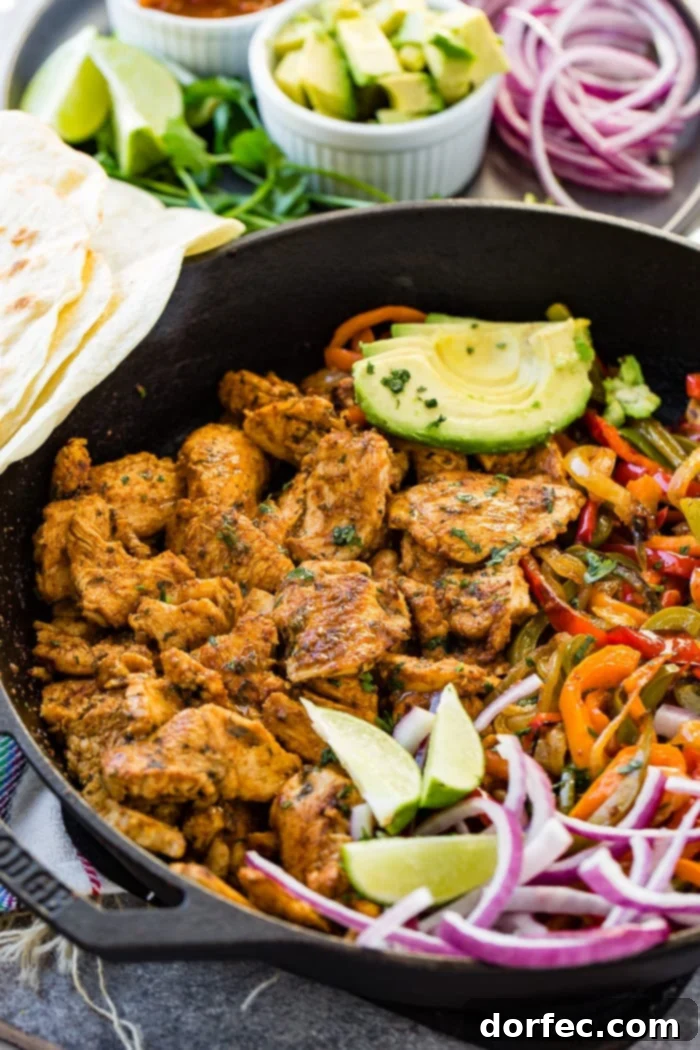 A photo of a cast iron skillet full of chicken fajita filling - cooked chicken, sauteed peppers and onions, lime wedges, sliced purple onion, and sliced avocados.