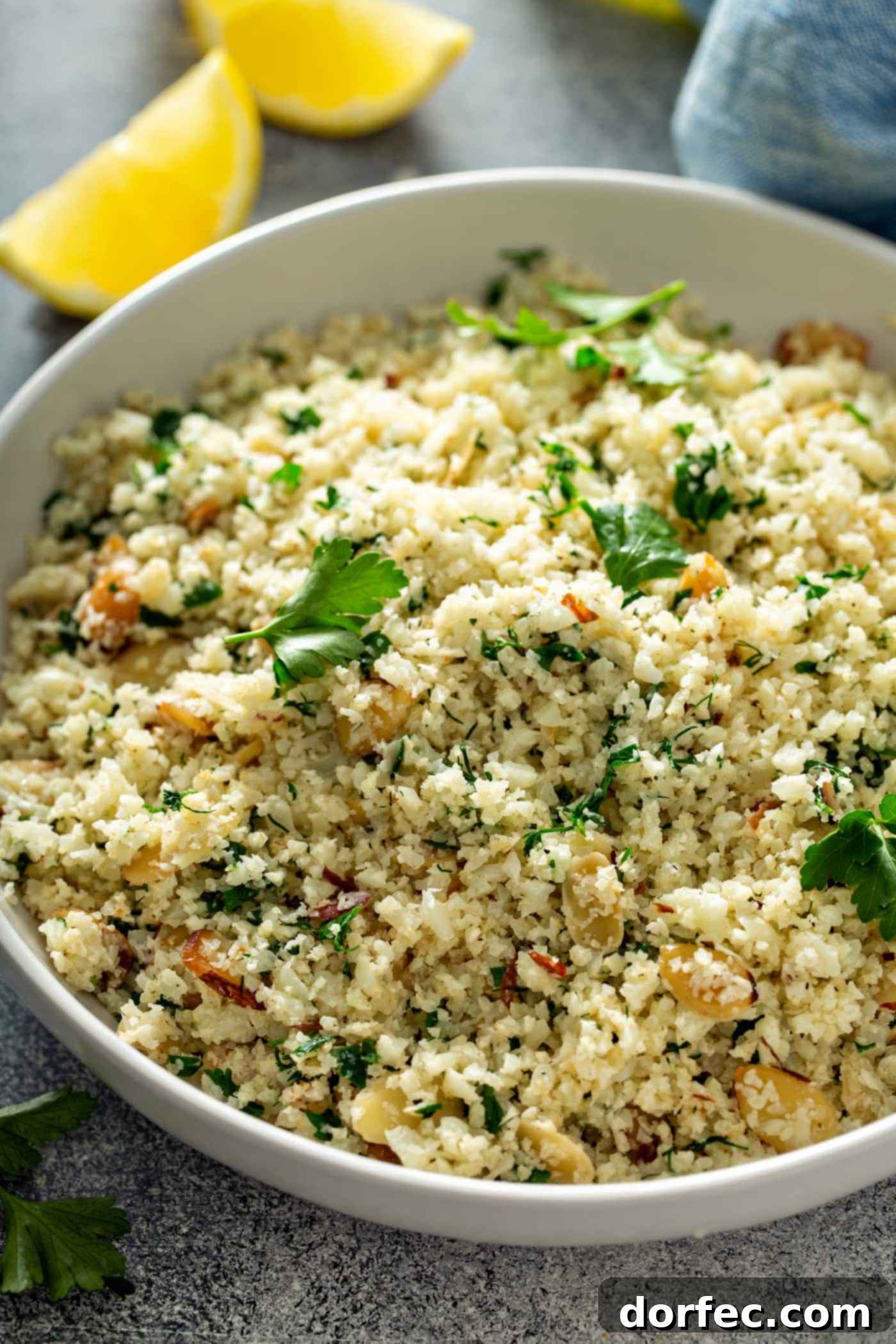 A close-up of a bowl of garlic herb cauliflower rice. There are green herbs, garlic, and sliced almonds in the rice, with fresh parsley leaves scattered on top. A lemon wedge is in the background.