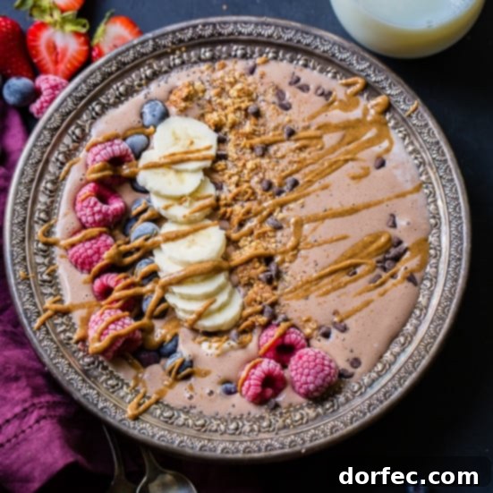 Kid Friendly Chocolate Banana Smoothie Bowl: A vintage silver bowl filled with a chocolate smoothie, topped with raspberries, bananas, blueberries, granola, and chocolate chips, drizzled with peanut butter. A purple napkin and glass of milk are in the background.