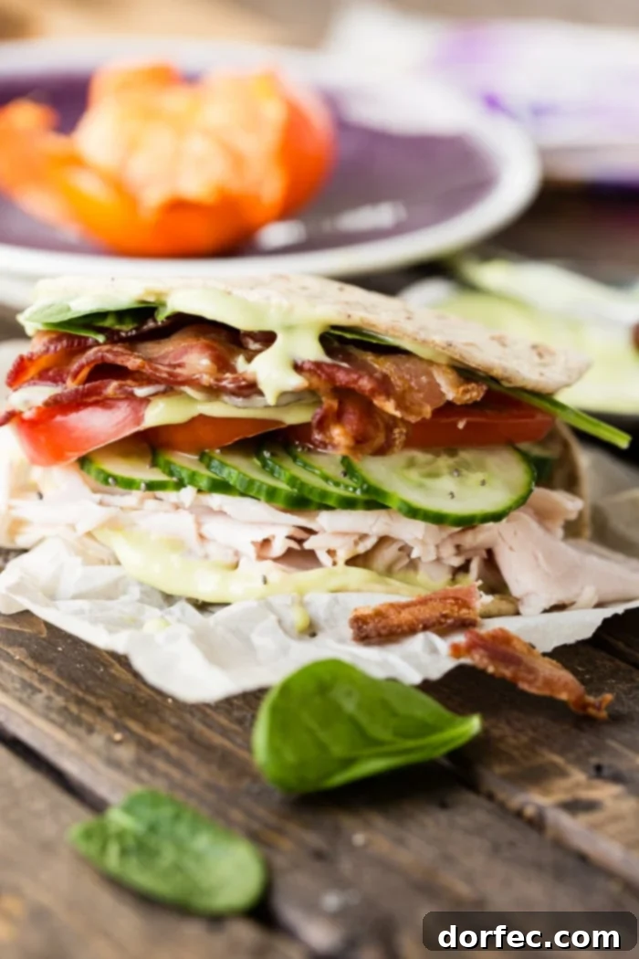 Flatbread sandwich sitting on parchment paper full of turkey, bacon, avocado dressing, cucumbers, tomatoes and chia seeds with a little spinach. Wood table with a piece of spinach on it and a cutie orange on a purple plate in the background next to avocado dressing.