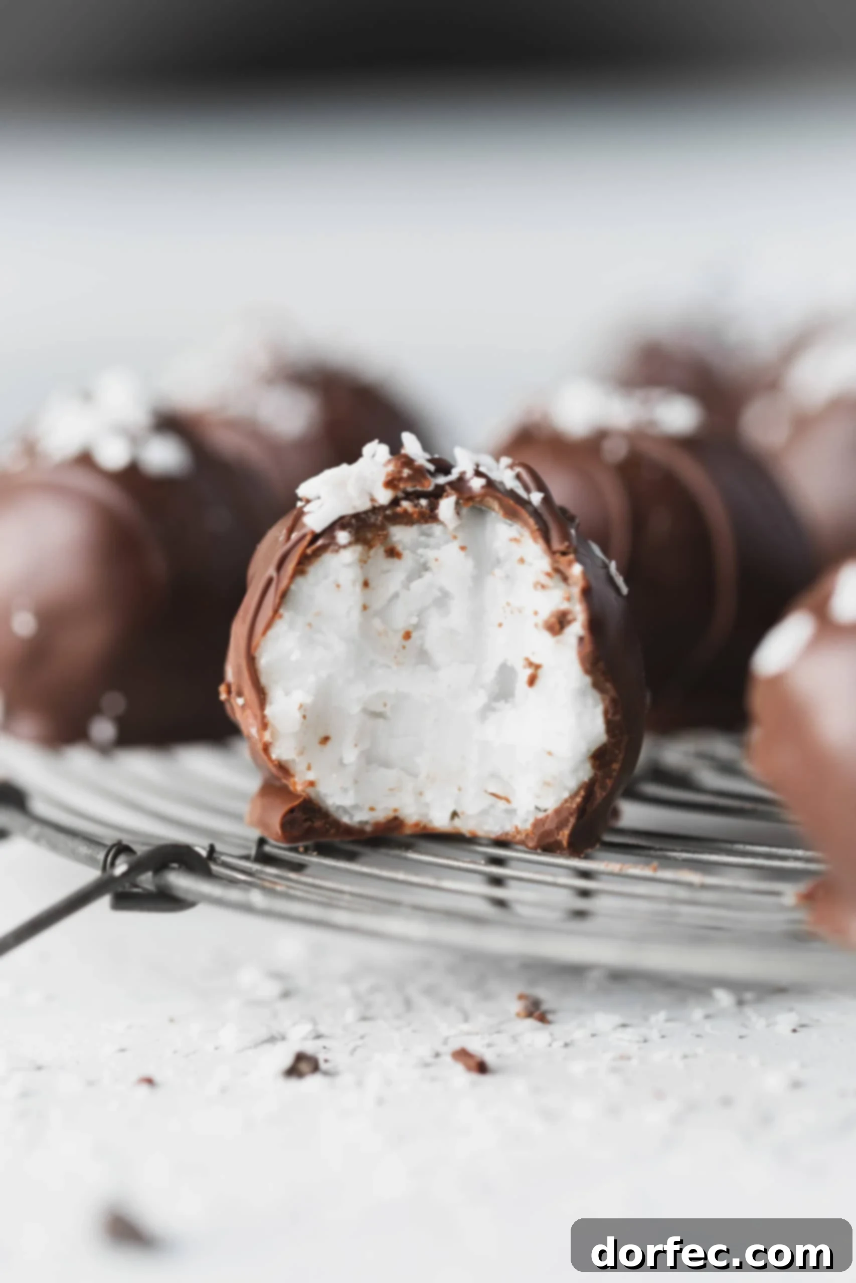 A chocolate covered coconut ball broken in half, revealing its creamy white coconut center, with other coconut balls blurred in the background. Perfect for a holiday treat!