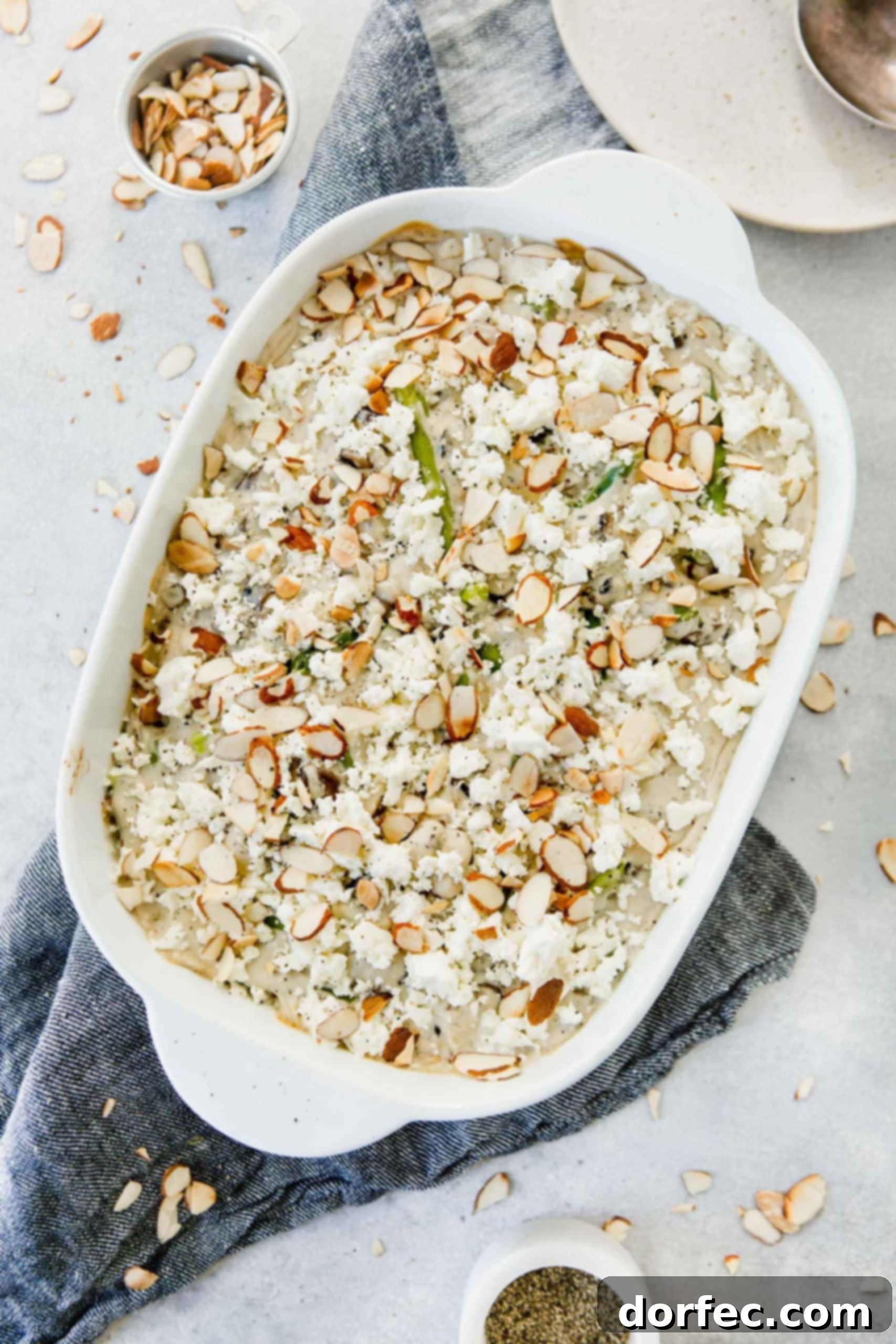 Feta Green Bean Casserole A close-up shot of the Feta Green Bean Casserole, showcasing the creamy white sauce enveloping the green beans, with generous crumbles of feta cheese and toasted sliced almonds scattered on top. A serving spoon is in the dish.