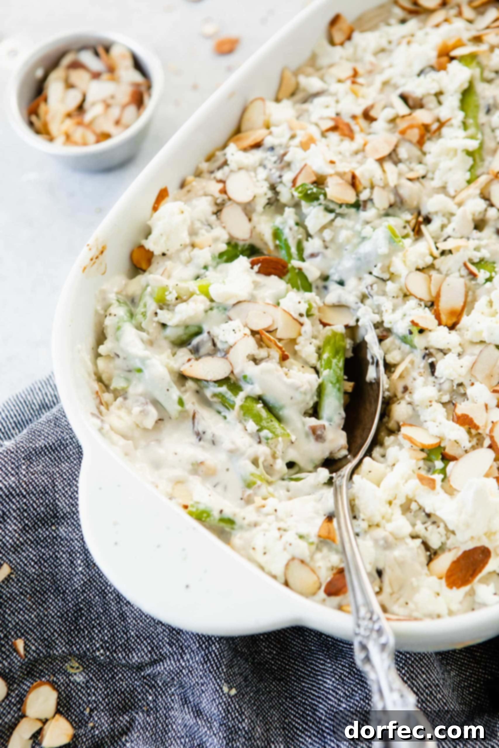 Feta Green Bean Casserole A close-up picture of Feta Green Bean Casserole in a serving dish, highlighting the tender green beans coated in a creamy white sauce, generously topped with crumbled feta cheese and golden-brown sliced almonds. A serving spoon is in the dish.