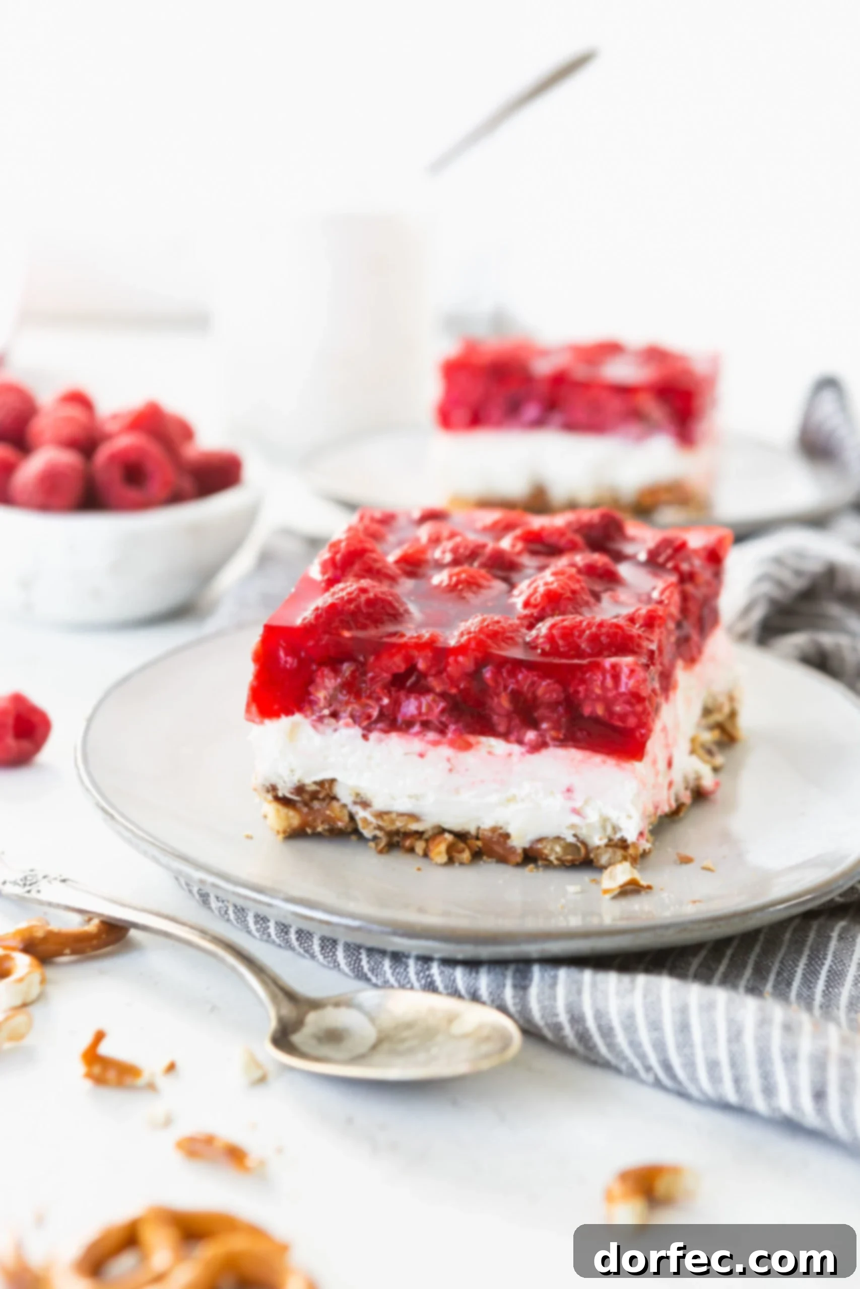 A close-up of a single serving of Raspberry Pretzel Jello Salad on a white plate, highlighting the perfectly defined layers: a crunchy pretzel base, a smooth white cream cheese layer, and a clear red jello layer studded with whole fresh raspberries. Other servings are blurred in the background.