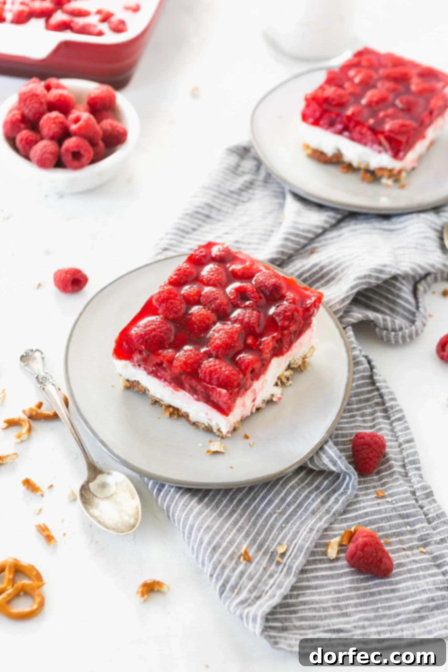 A close-up of a serving of Raspberry Pretzel Jello Salad on a white plate, emphasizing the crispy pretzel crust, the creamy white filling, and the ruby-red jello layer topped with whole fresh raspberries. A spoon, a bowl of fresh raspberries, and another serving are in the background.