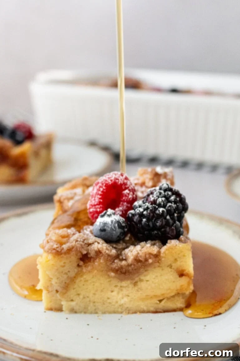 Overnight French Toast Bake 12 A hand is gently pouring golden syrup over a piece of French toast casserole, beautifully garnished with fresh berries and a dusting of powdered sugar.