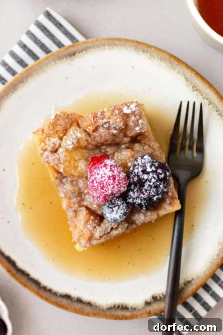 Overnight French Toast Bake 13 An inviting top-down shot of a piece of French toast casserole, bathed in a pool of golden syrup, adorned with an assortment of fresh berries and a delicate dusting of powdered sugar.