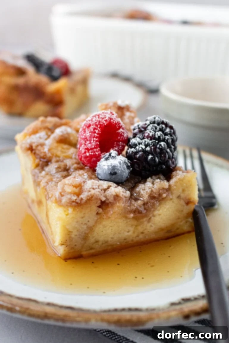 Overnight French Toast Bake 3 A close-up of a delectable slice of French Toast Casserole, drizzled with golden maple syrup and topped with a medley of fresh berries, resting on a plate with a fork beside it.
