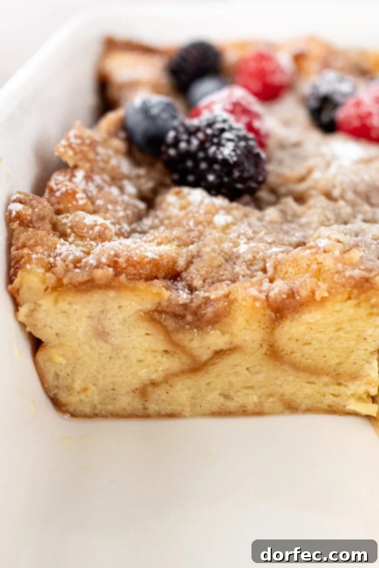 Overnight French Toast Bake 4 A perfectly baked slice of Easy French Toast Casserole, adorned with a delicate dusting of powdered sugar and a vibrant assortment of fresh berries, is carefully lifted from a full baking dish.
