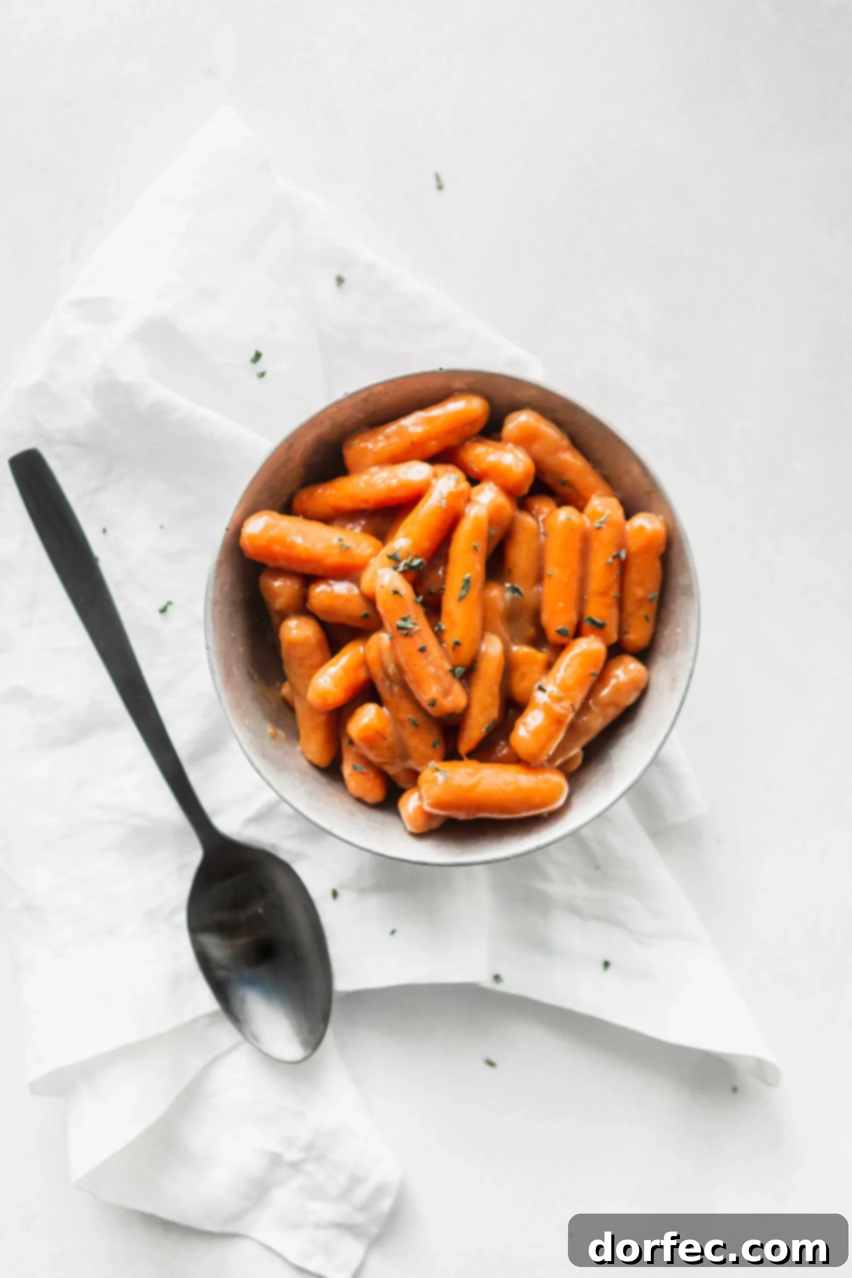 A serving bowl of orange glazed baby carrots. There are a few herbs sprinkled on top and a serving spoon is next to the bowl.