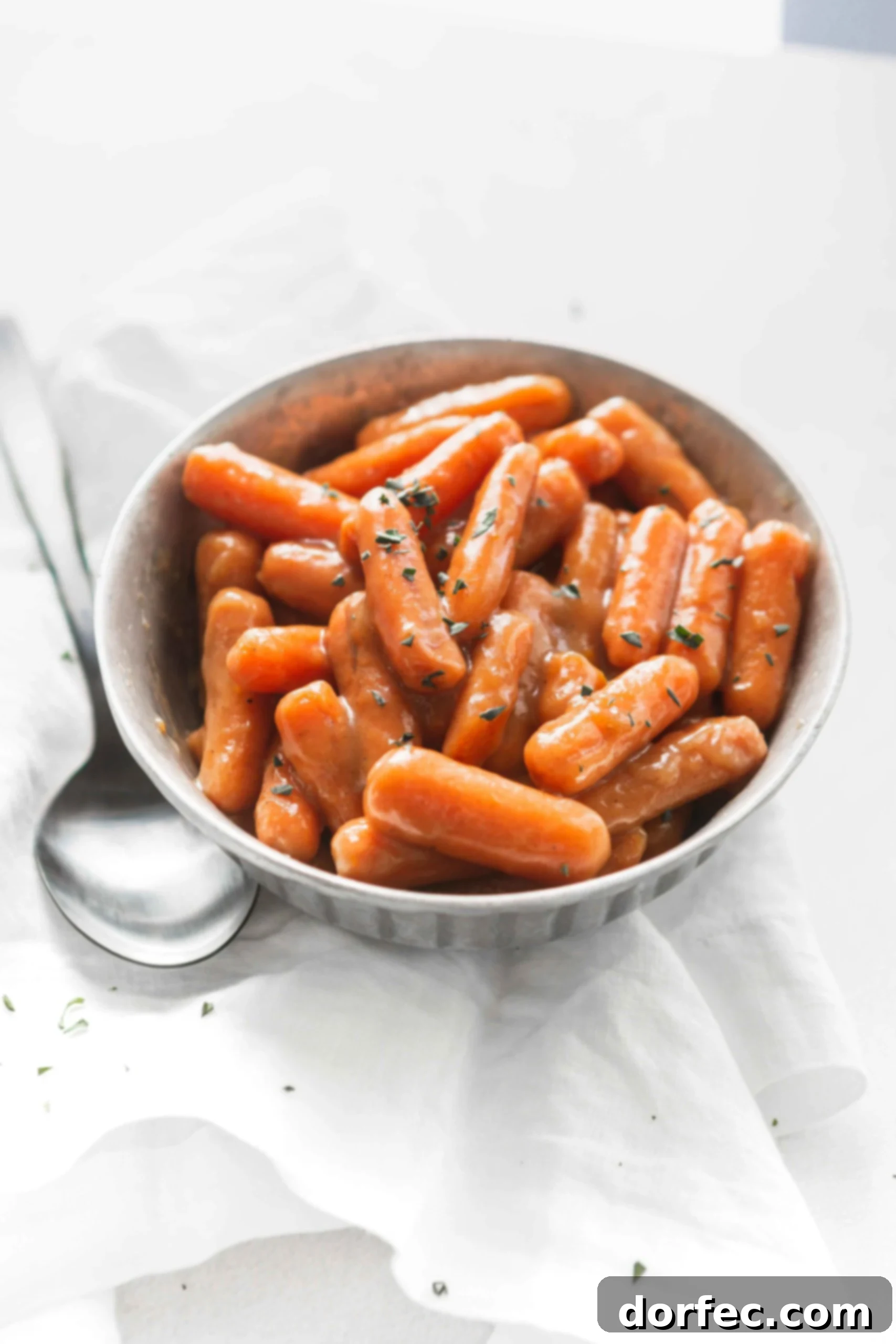 A bowl of orange glazed baby carrots with a little parsley sprinkled on top. A serving spoon is next to the bowl of carrots.