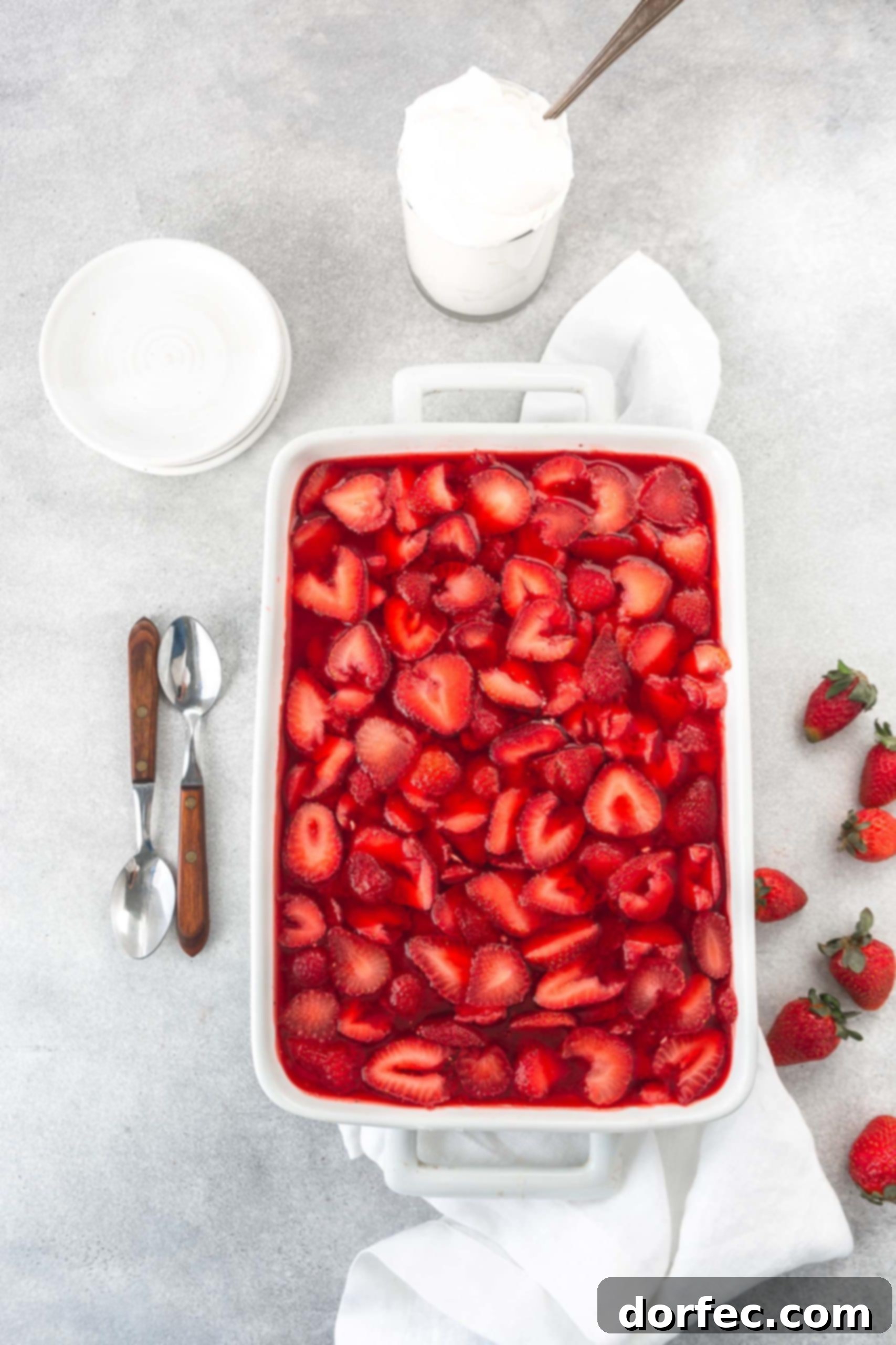 A casserole dish of strawberry pretzel jello salad. The strawberry jello is loaded with strawberry slices and there are two spoons and several strawberries beside the dish. A white bowl is in the background. The