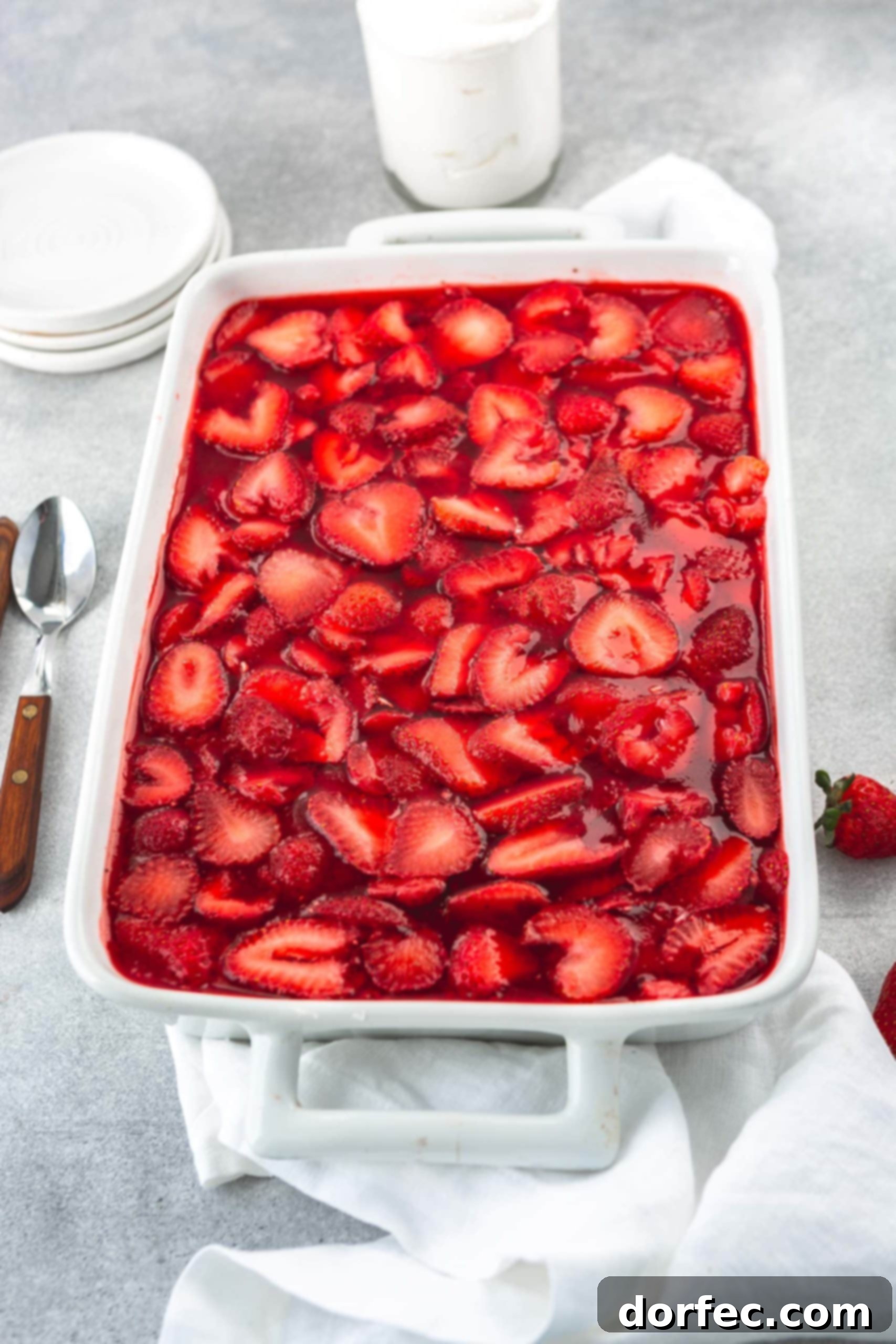 A white casserole dish with strawberry pretzel jello salad. The jello is filled with sliced strawberries and there are strawberries and two spoons next to the casserole dish.