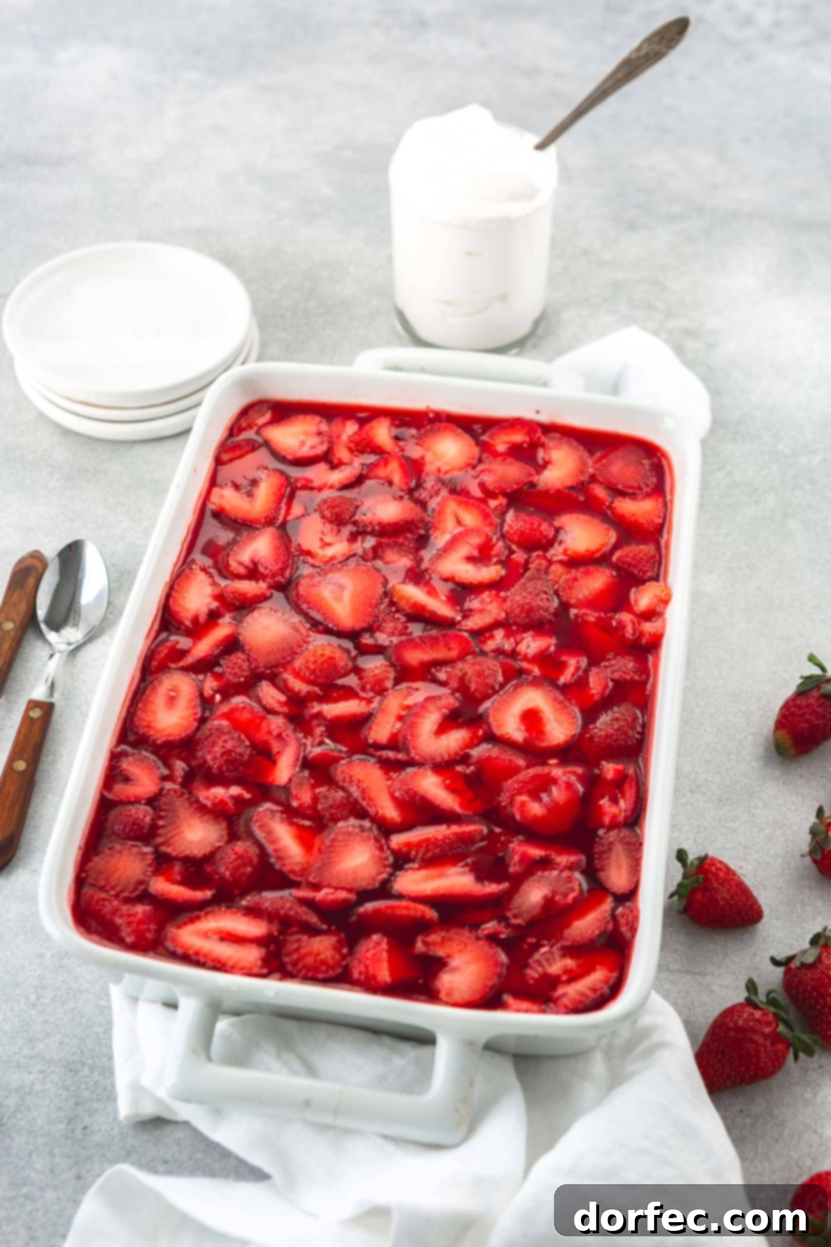 A white casserole dish with strawberry pretzel jello salad. The strawberry jello is filled with sliced strawberries. Two spoons, strawberries and a couple of small white bowls are in the background.