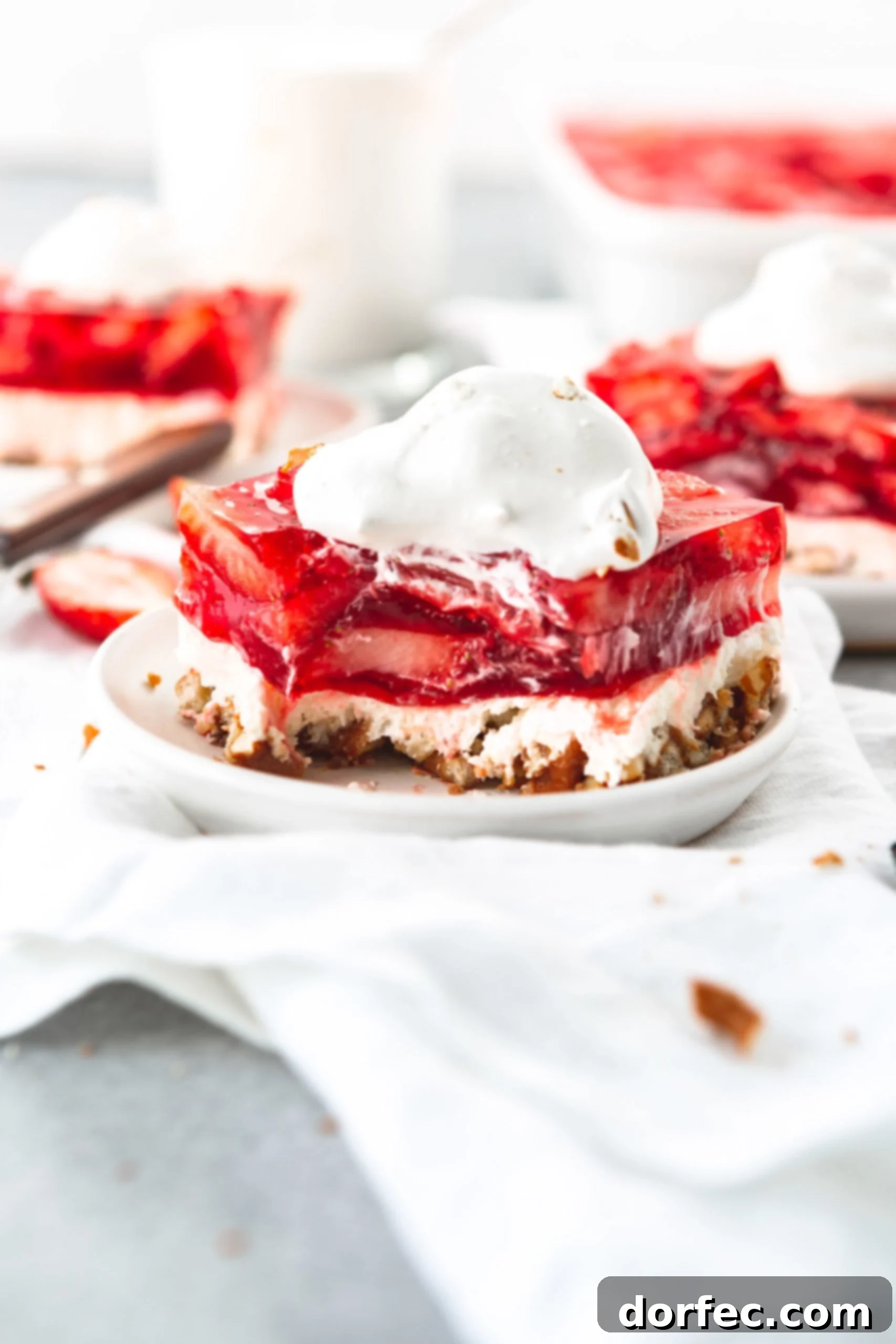 A serving of strawberry pretzel jello salad on a white salad plate. The salad has three layers, pretzel on the bottom, a cream layer in the middle and strawberry jello with sliced strawberries on top. The salad is topped with a dollop of cool whip. There are two other servings in the background.