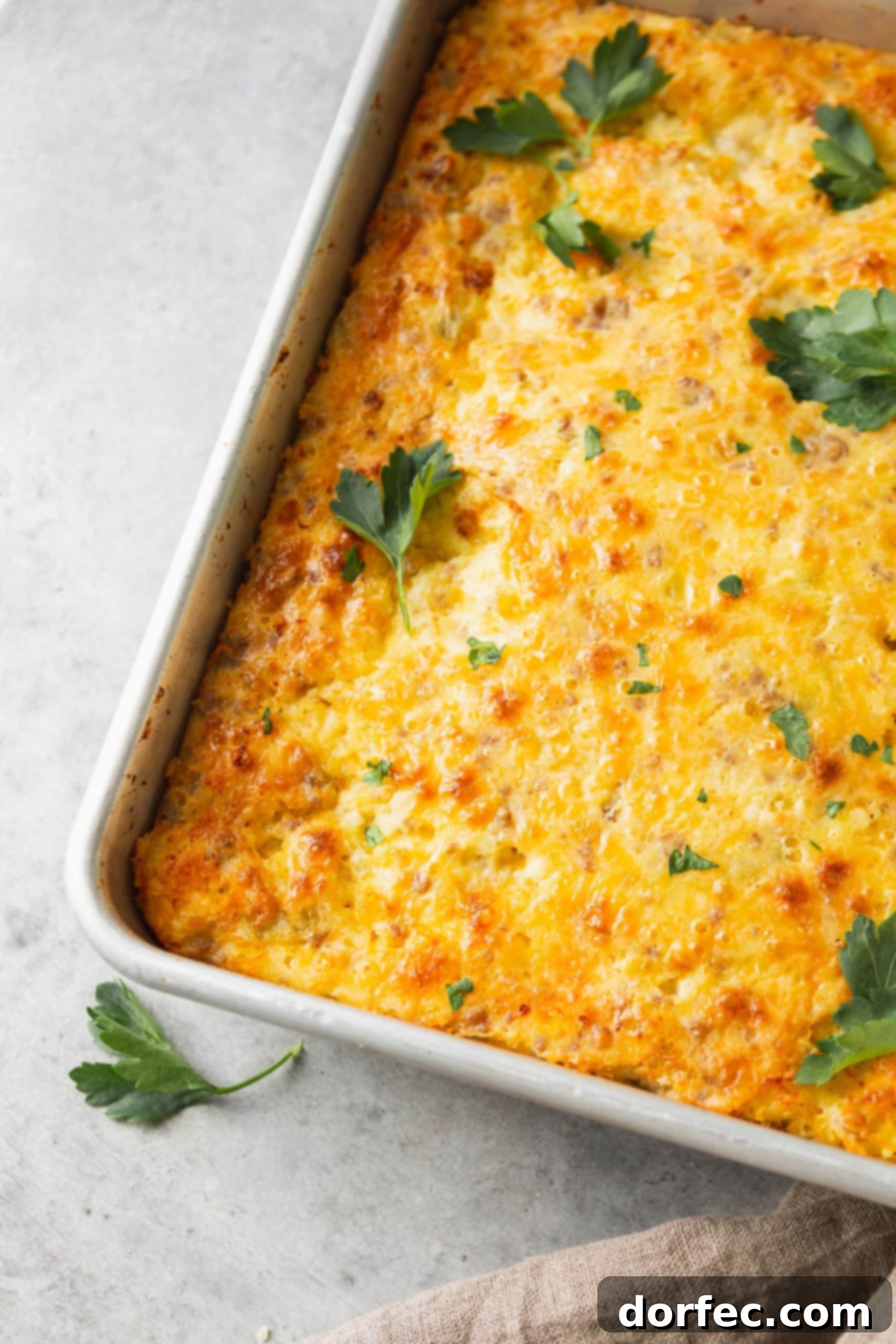 A casserole dish of sausage, cheese soufflé, adorned with a few scattered parsley leaves for garnish, showcasing its yellow color with bits of sausage and melted cheese.